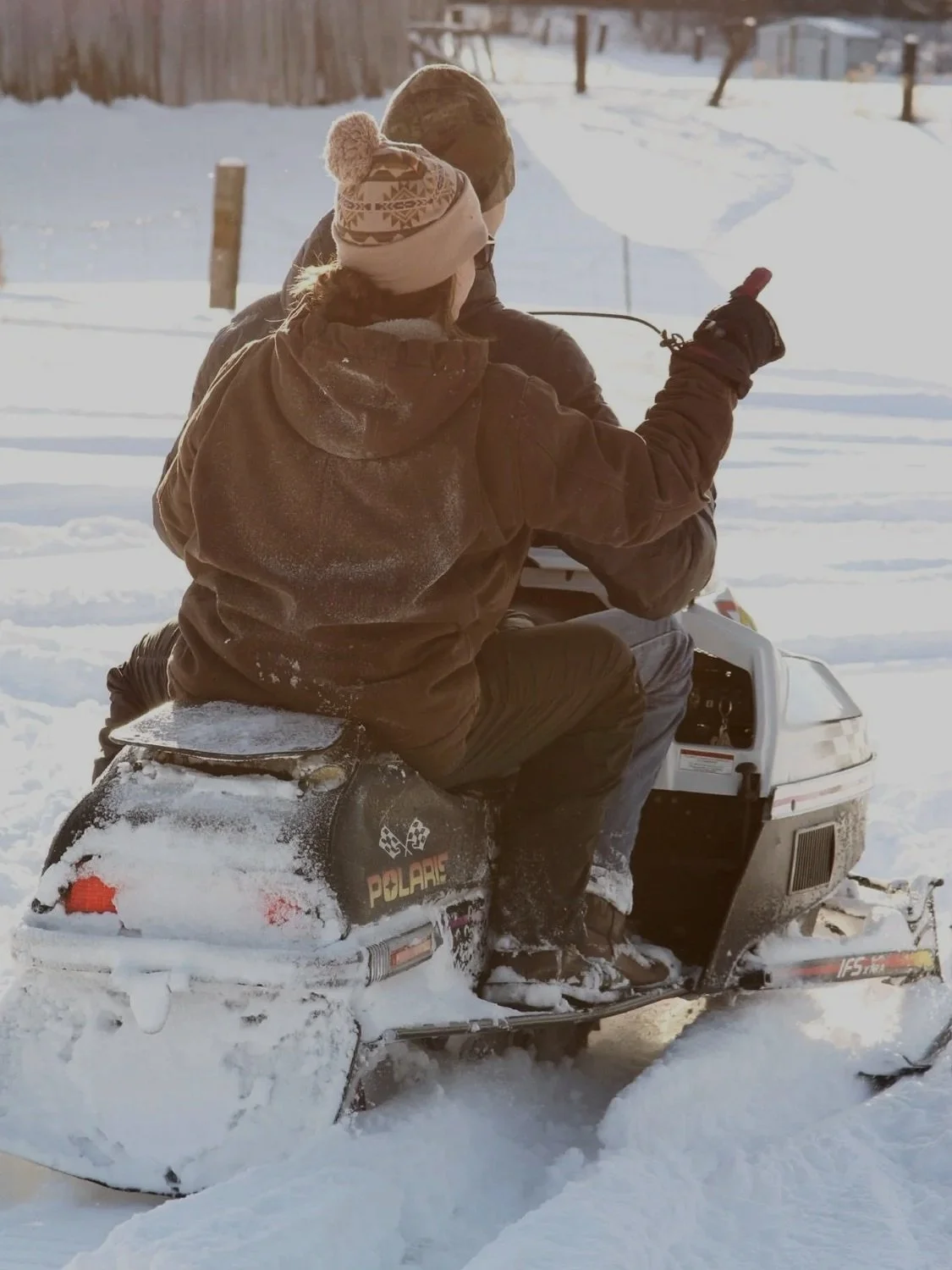 Two people in winter clothing riding a snowmobile through snow-covered terrain, with one person giving a thumbs-up.