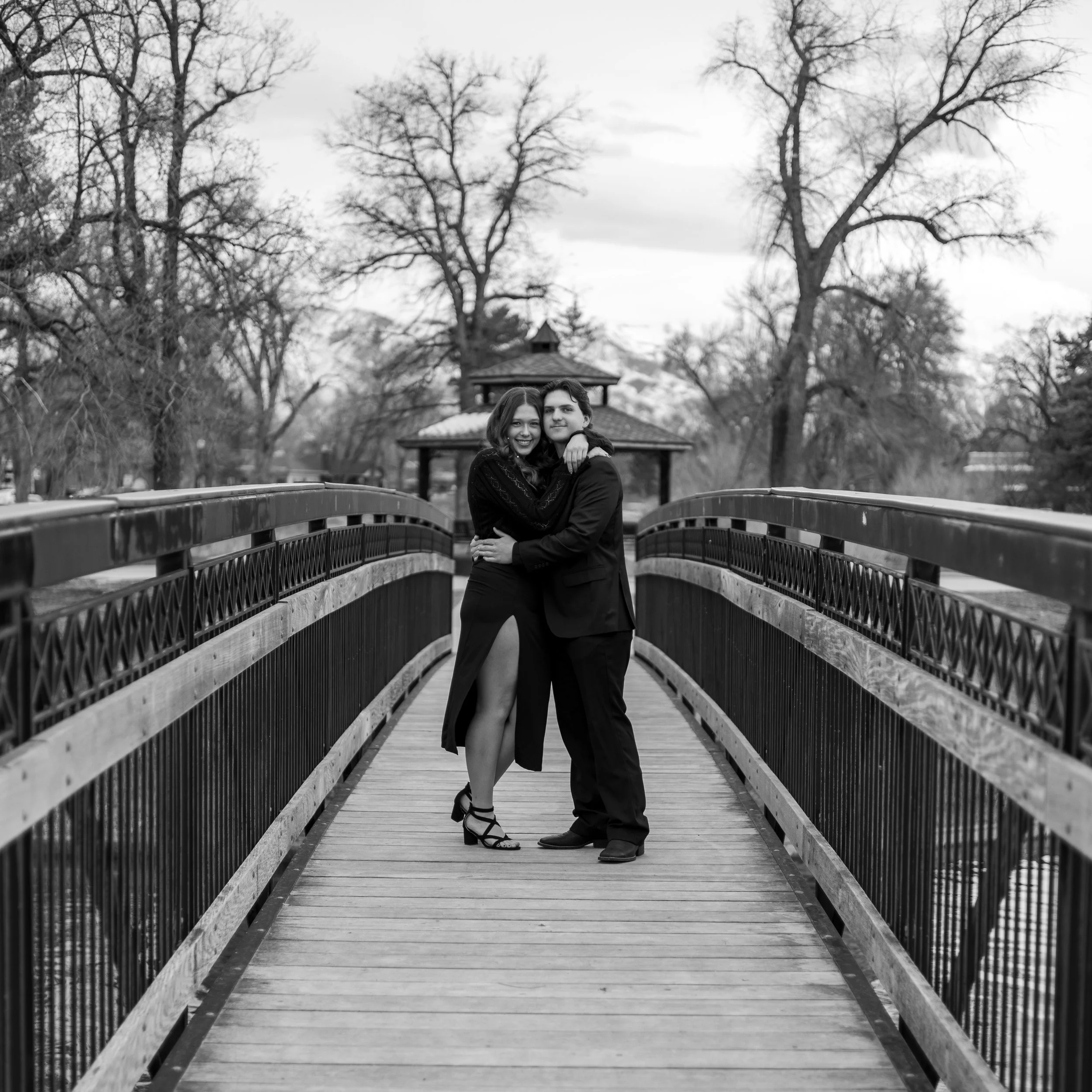 A black and white photo of a couple standing on a bridge, embracing each other and smiling, with bare trees and a gazebo in the background.