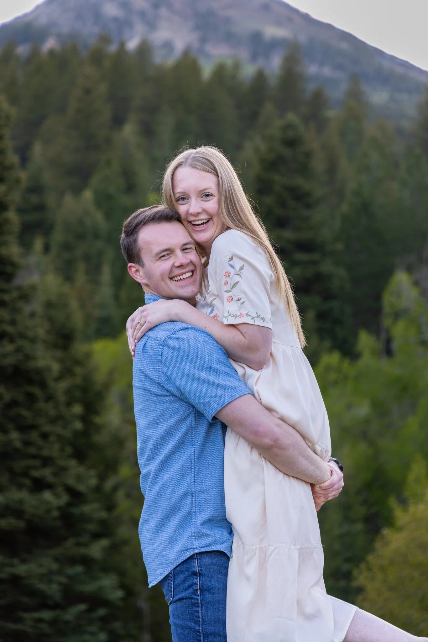 A man holding a woman in a wooded outdoor setting with mountains in the background, both smiling and happy.