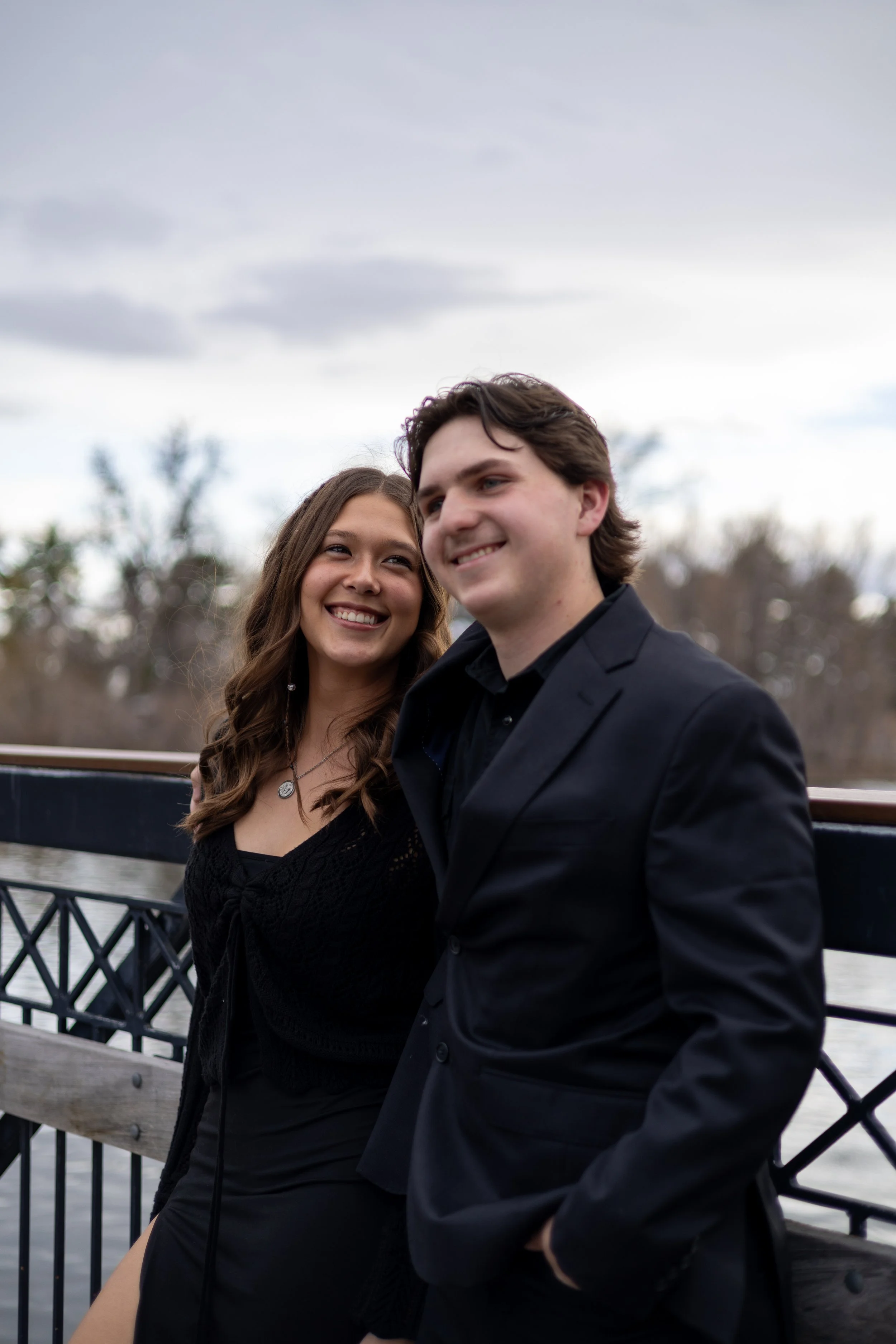 A smiling young woman and man in formal attire standing on a bridge outdoors with trees and cloudy sky in the background.
