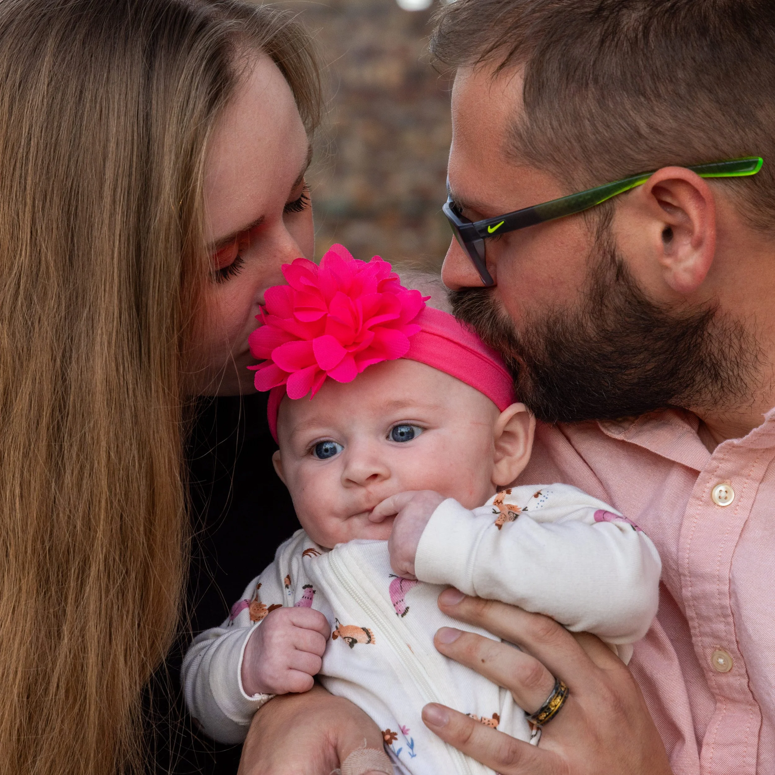 A baby girl with a large pink flower headband being kissed by her father with glasses and a beard and her mother with long brown hair, against a blurred outdoor background. Taken by Tucker Davis at the American Fork, Utah, amphitheater.