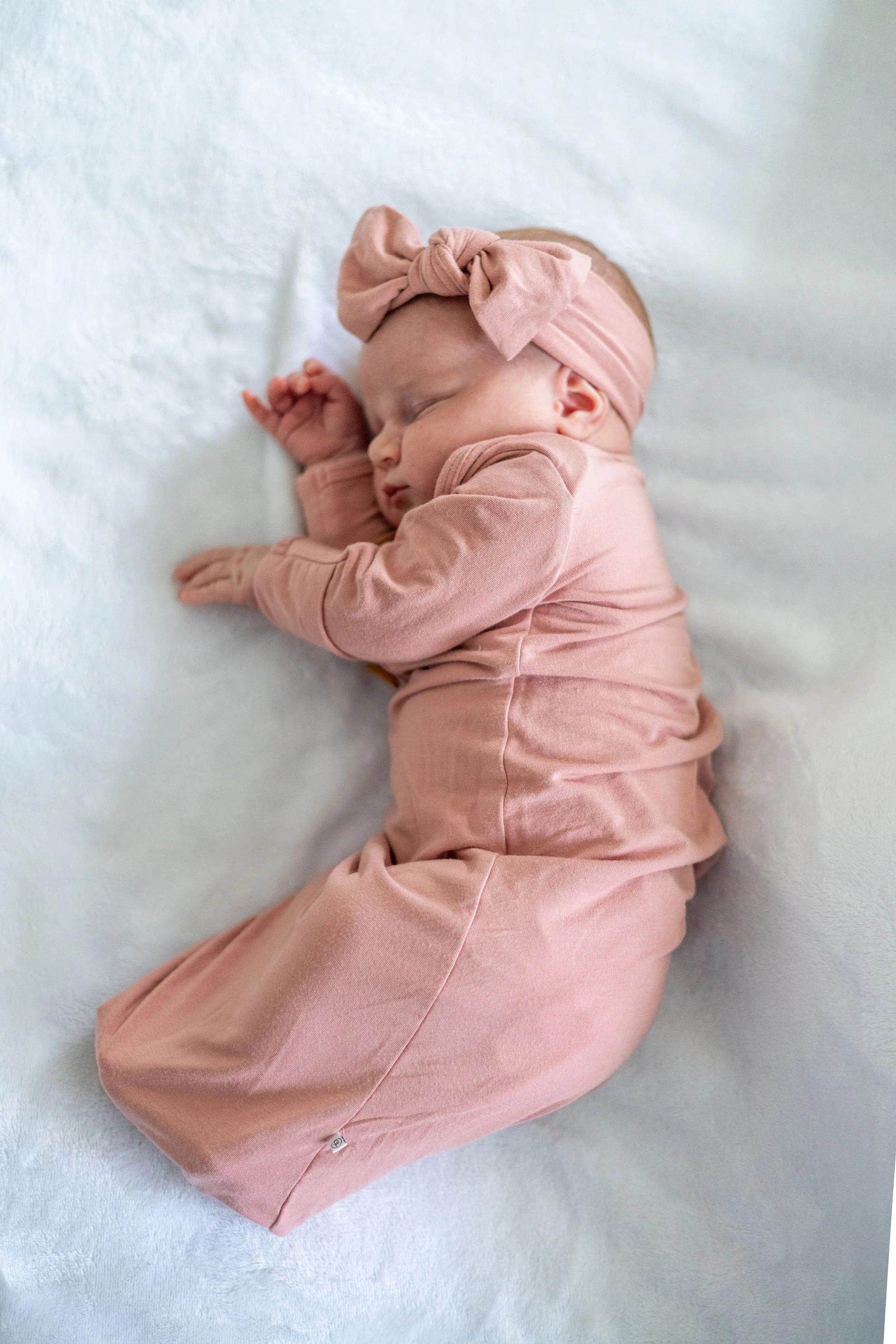 A sleeping baby girl wearing a pink outfit and a pink headband with a bow, lying on a soft, white surface.