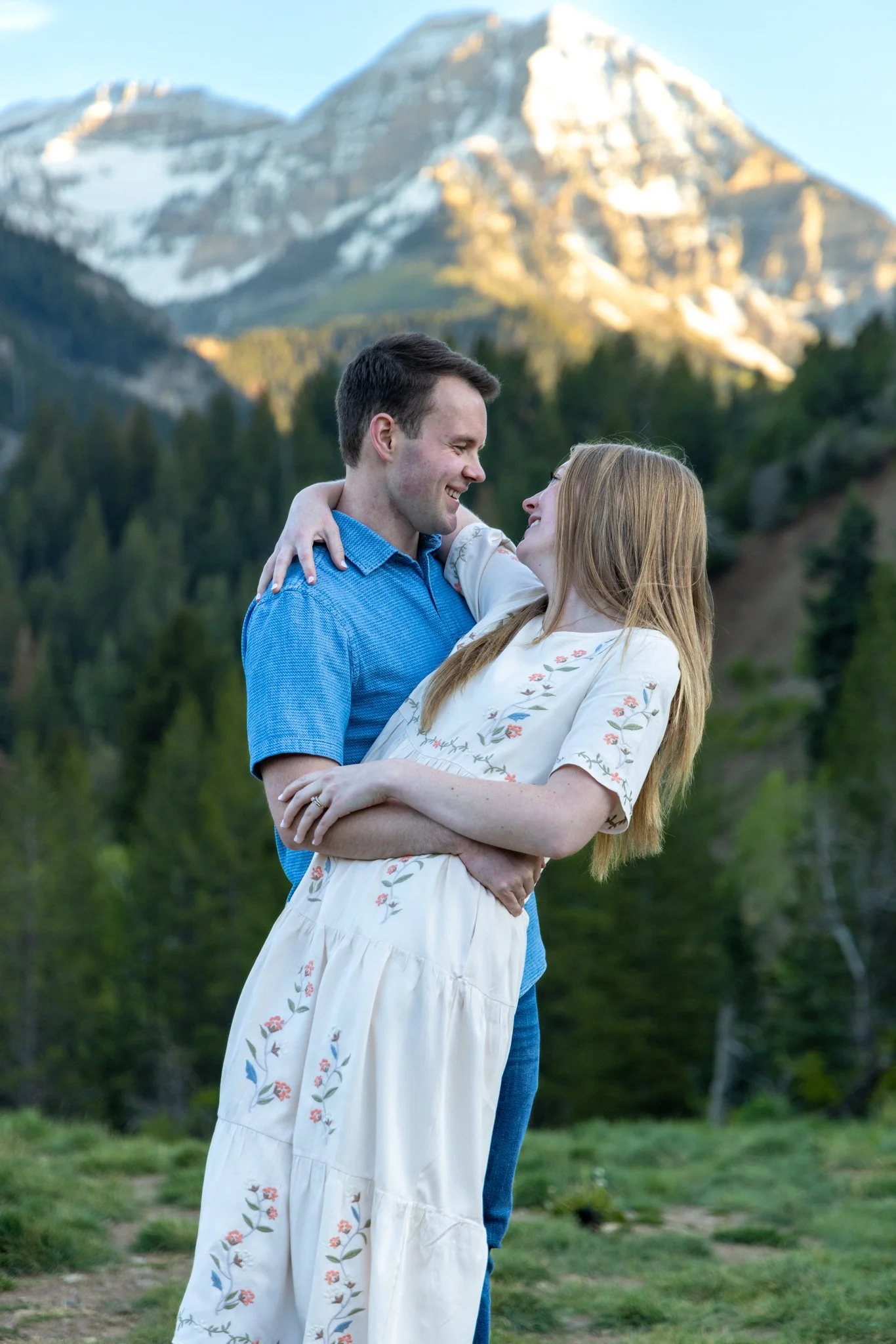 A young man and woman holding each other and smiling in a scenic outdoor setting with mountains and trees in the background.
