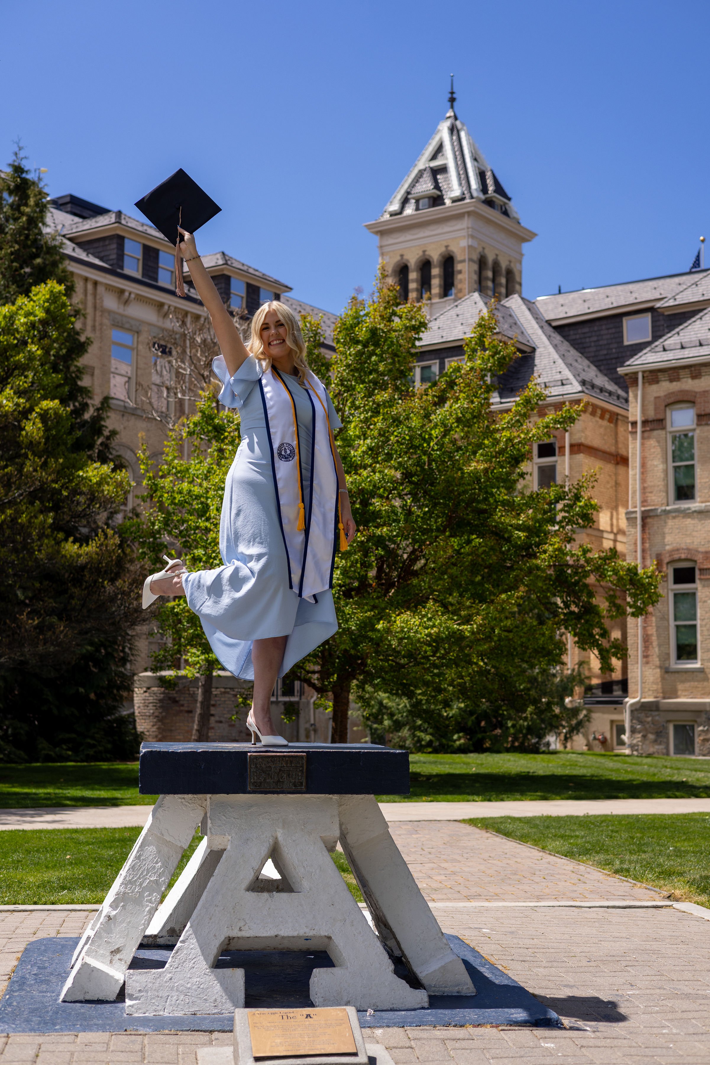 A woman in graduation attire, including a white gown and cap, is celebrating with her left arm raised, holding her cap, on a monument in a park with trees and historic buildings in the background on a sunny day.