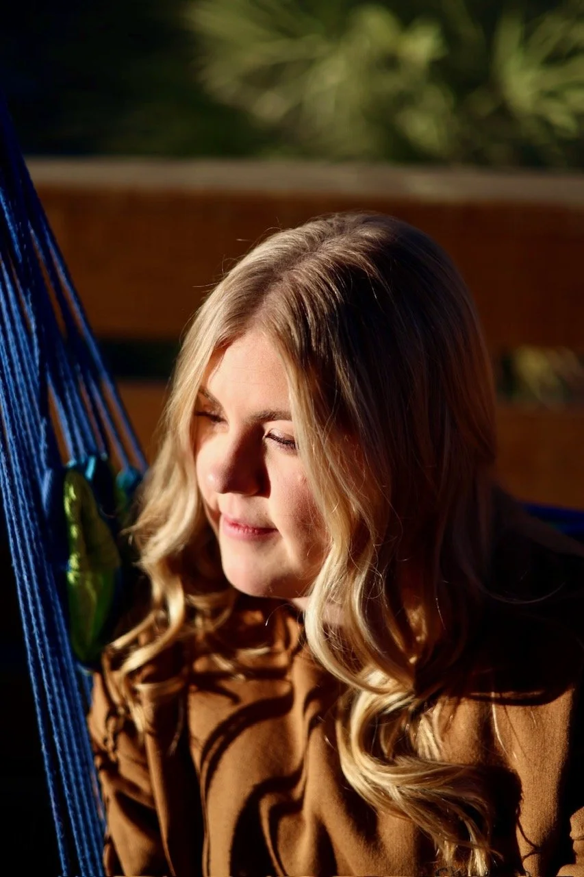 Close-up of a woman with blonde hair and closed eyes, relaxing in a blue hammock outdoors with greenery in the background.