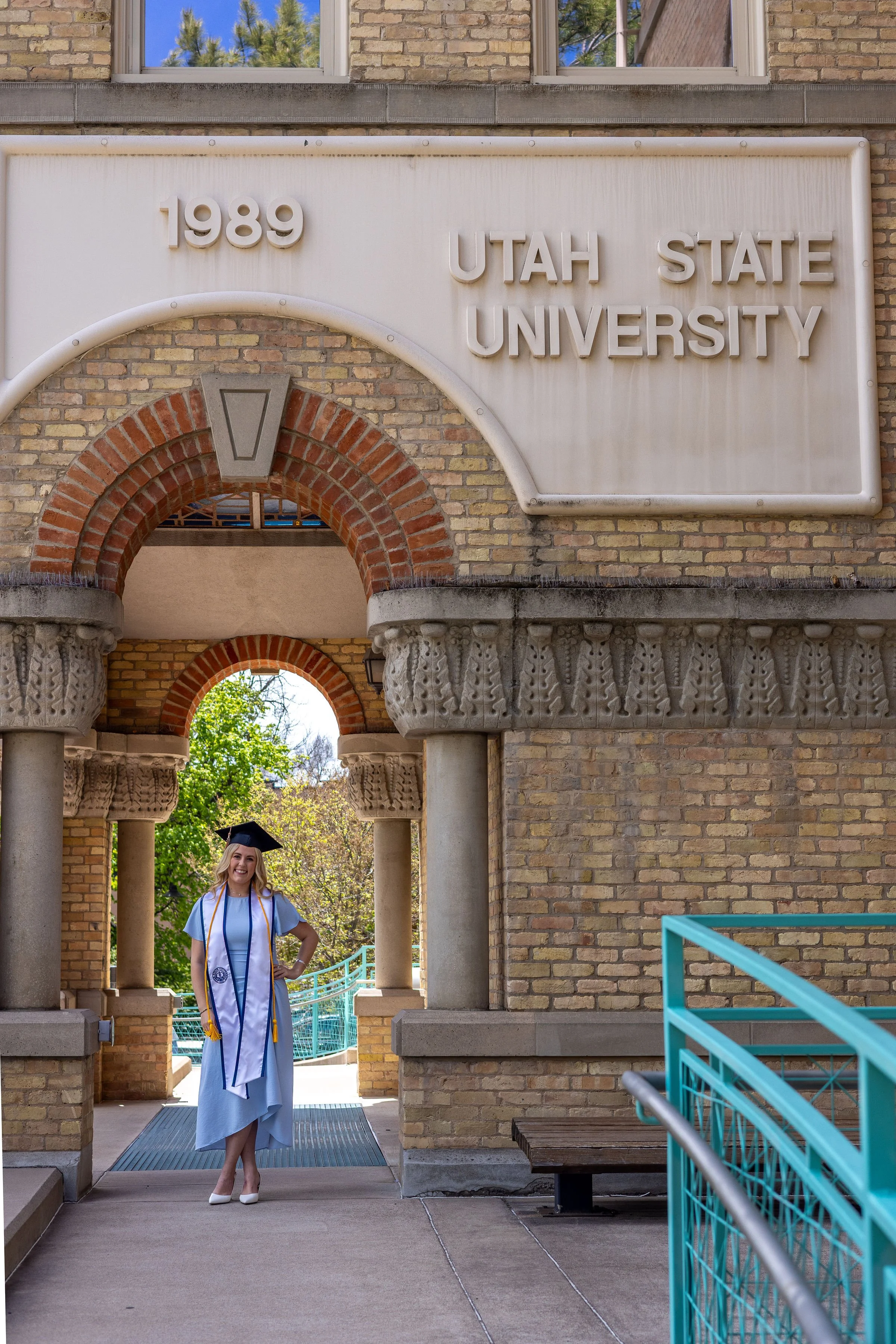 A smiling woman in a graduation cap and gown standing under a brick archway at Utah State University, with the university sign showing the year 1989 above her.