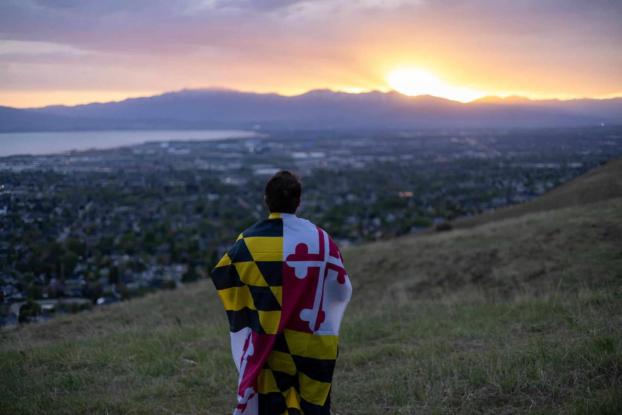 A man wrapped in a Maryland flag gazes at a sunrise or sunset over a city with mountains and a body of water in the background.