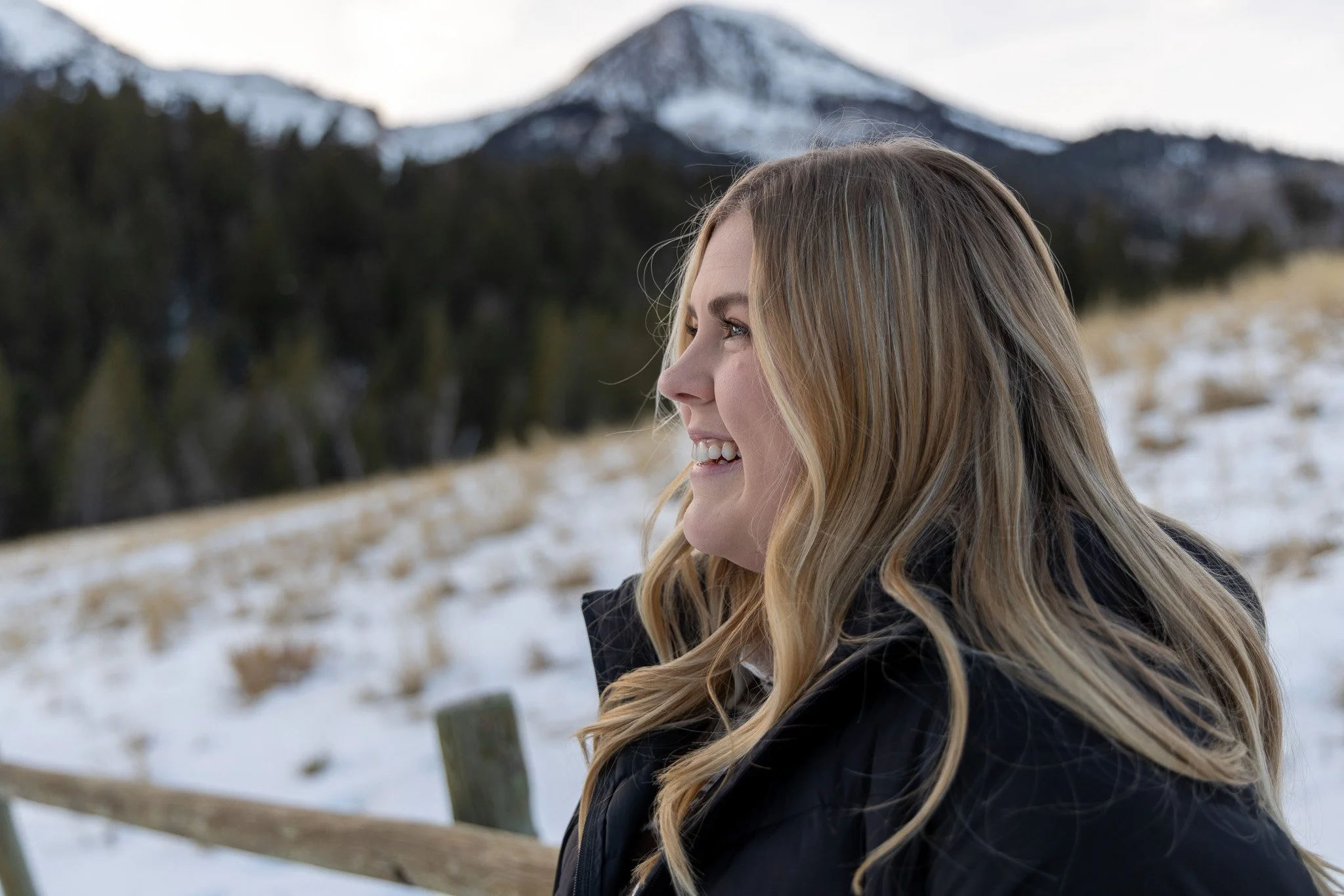 A woman with long blonde hair smiling outdoors in a snowy landscape with mountains and trees in the background.