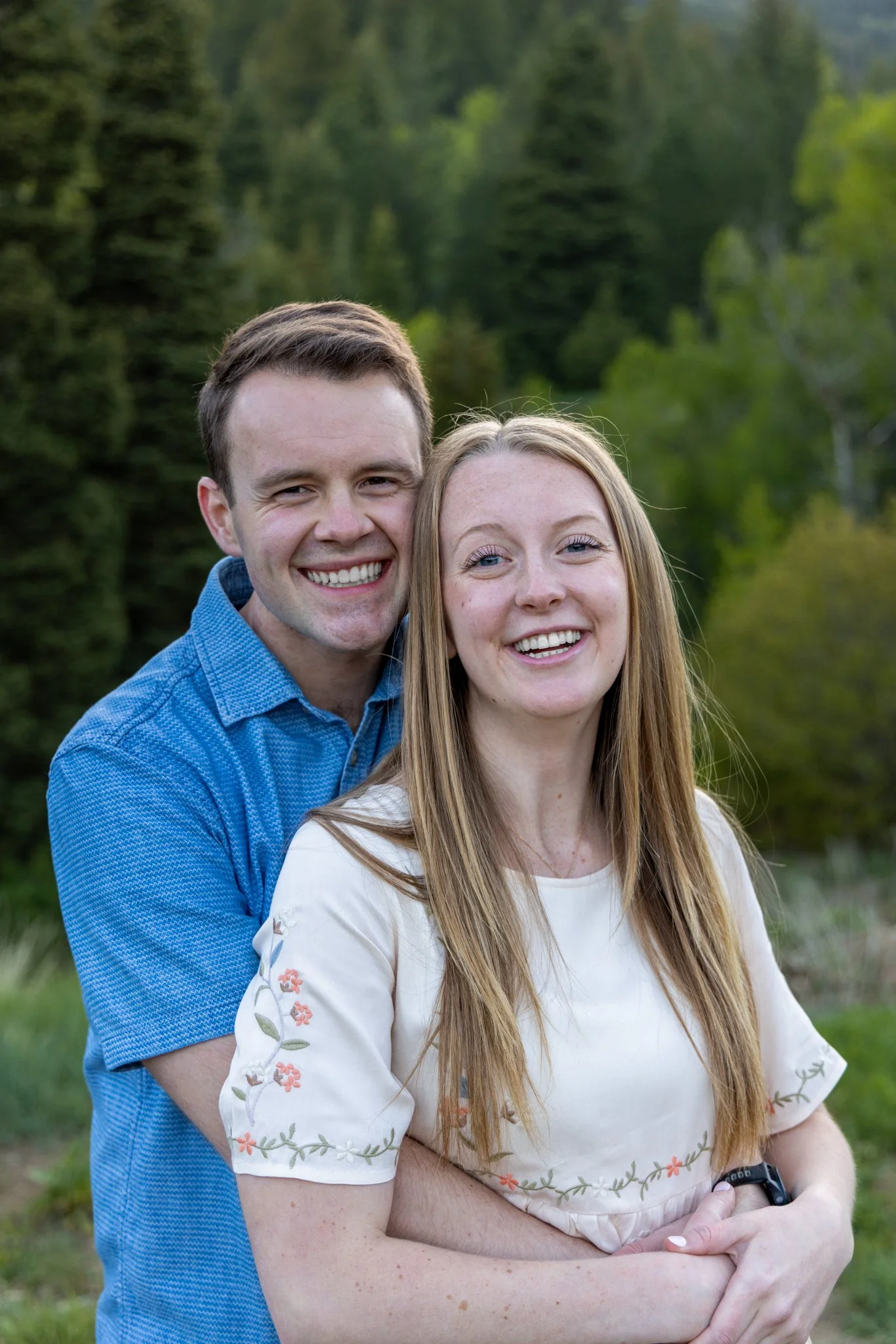 A smiling couple stands outside in front of a lush green forest, with the man standing behind the woman, wrapping his arms around her waist.