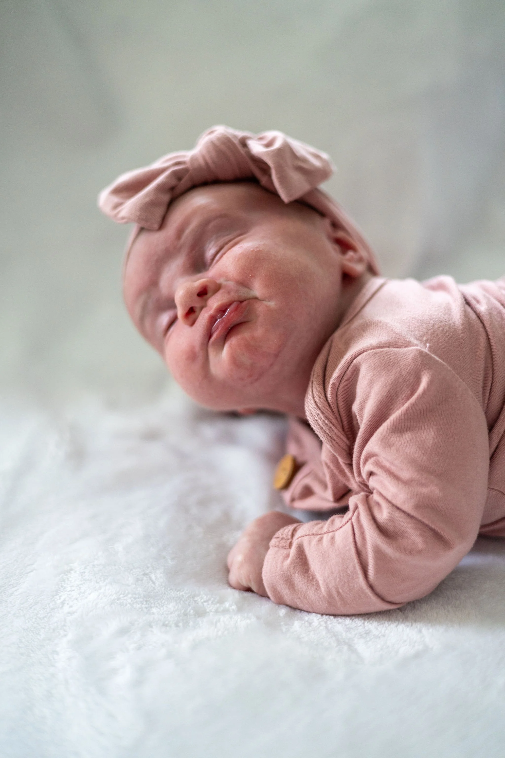 A sleeping baby wearing a pink headband with a bow, lying on their side on a soft white surface, dressed in a pink long-sleeve shirt.