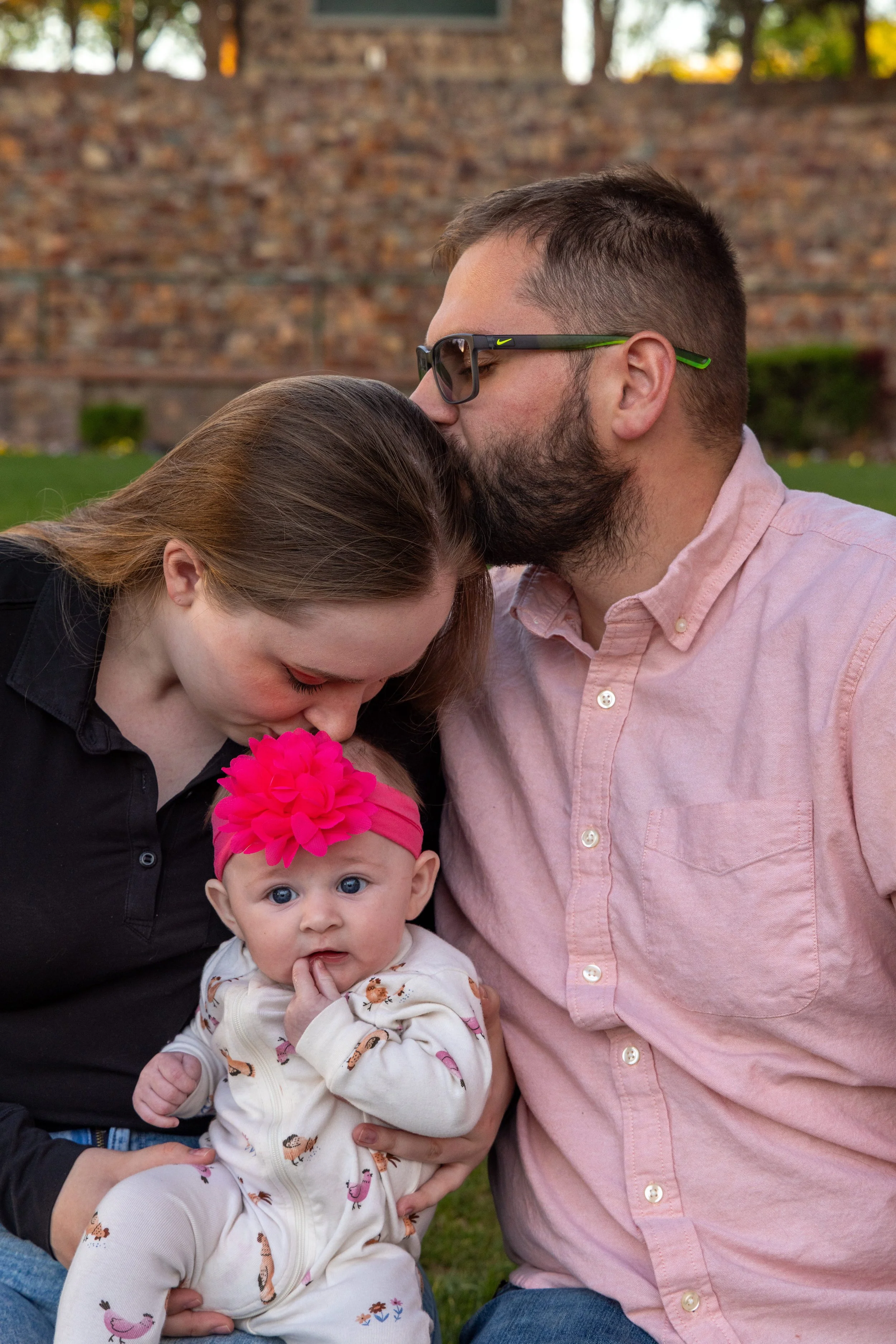 A family of three outdoors, with a woman and a man holding their baby girl. The woman leans her head down, and the man kisses her on the forehead. The baby girl has blue eyes, a pink headband with a large flower, and is in a white outfit with colorfu