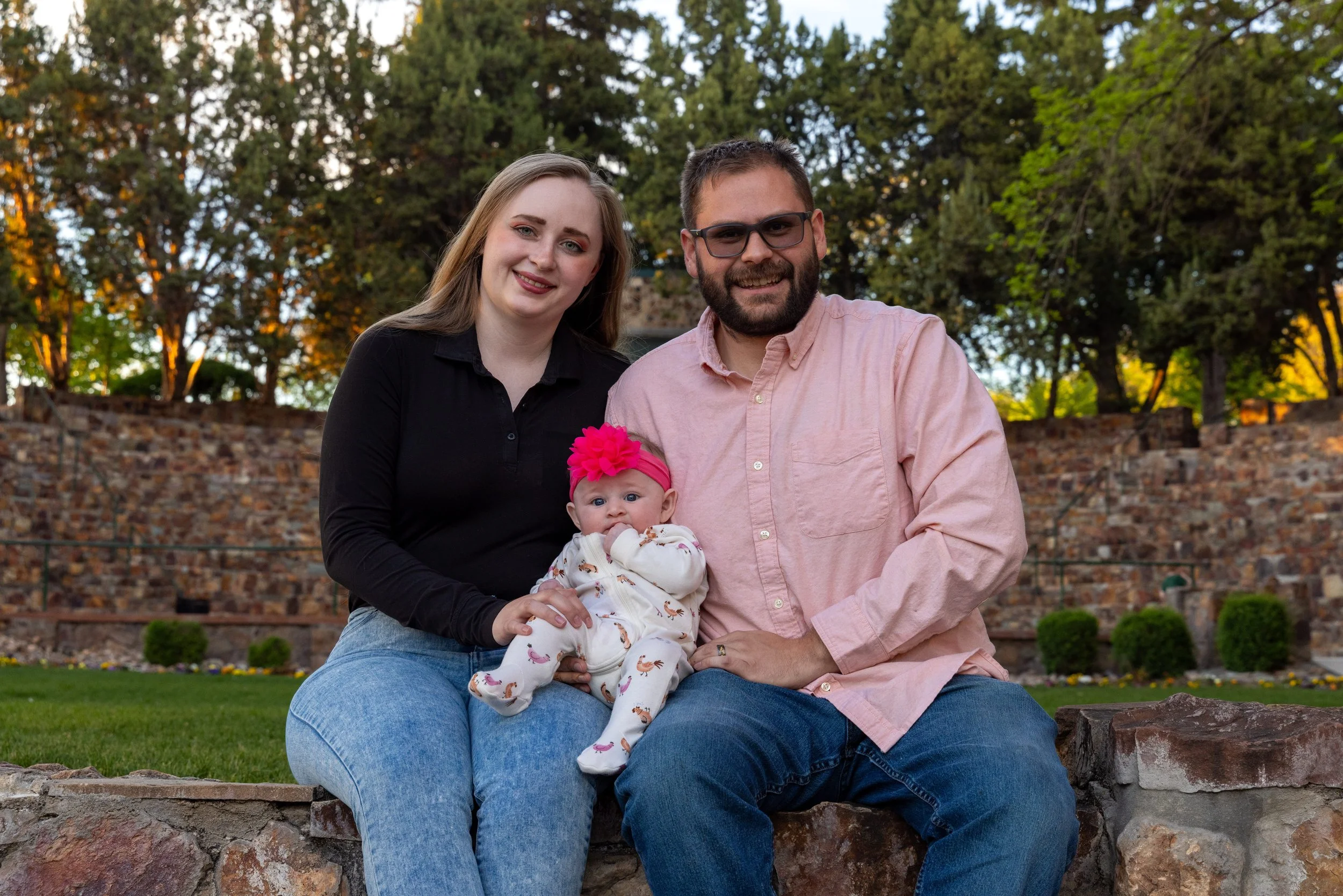 A family of three sitting outdoors on a stone wall in a park during sunset, with trees and a brick wall in the background. The mother has long brown hair, is wearing a black shirt and blue jeans. The father has a beard, is wearing glasses, a pink shi