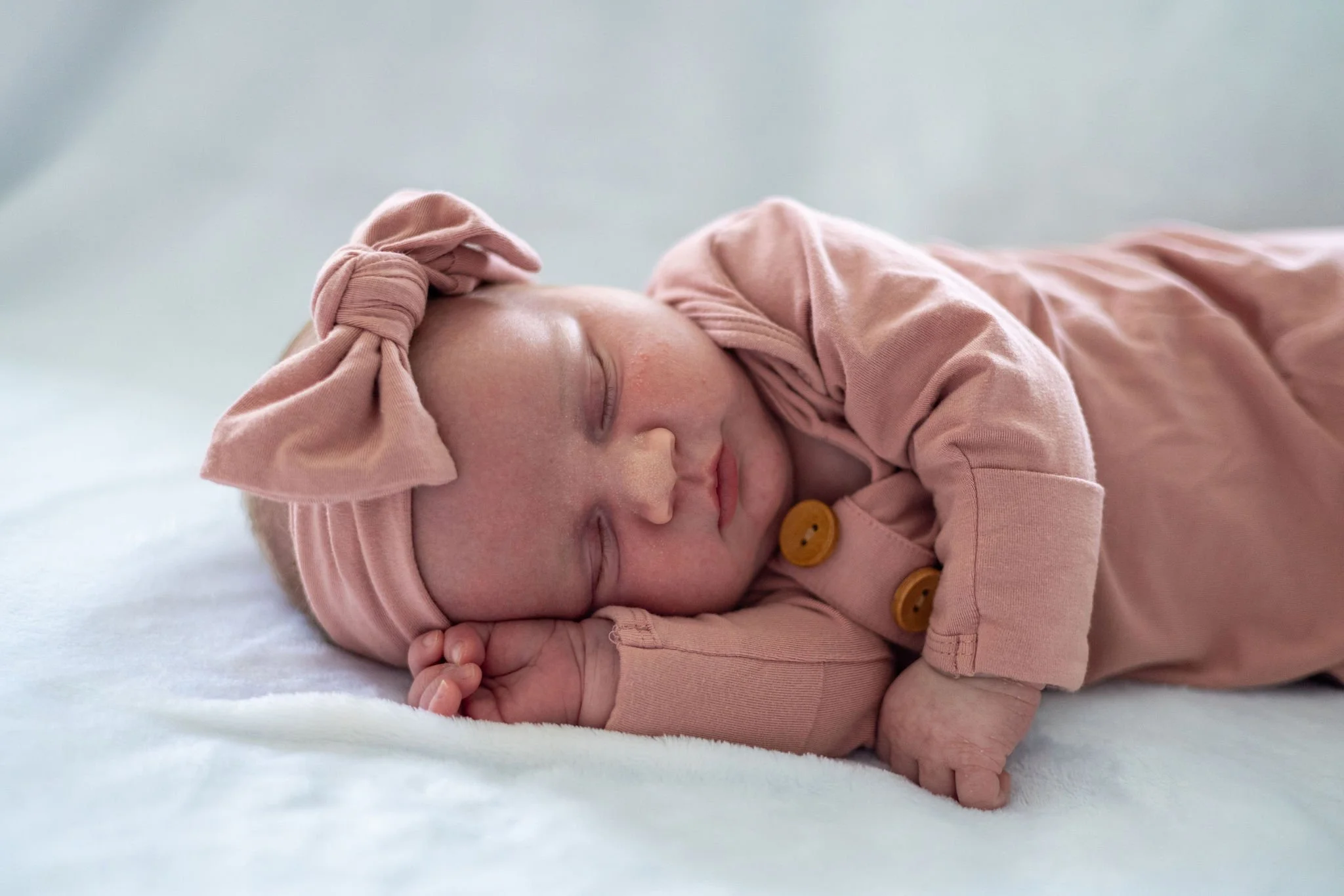 A sleeping baby girl dressed in a pink outfit and a matching pink headband, lying on her side on a white surface.
