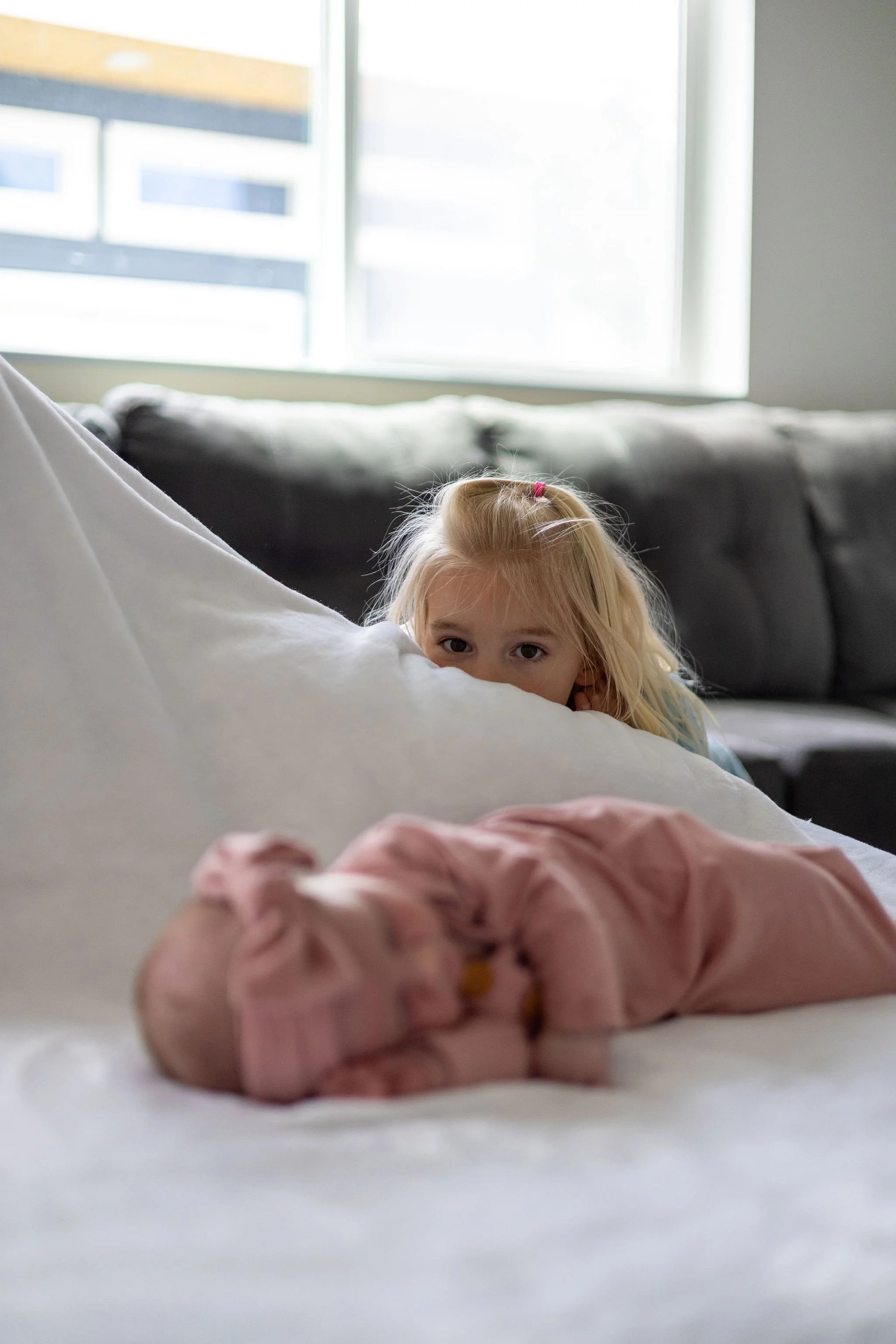 A young girl with blonde hair lying on a bed, partially hidden behind a large pillow, with her eyes peeking over the pillow. In the foreground, there is a pink scrunchy or hair accessory on the bed. The background shows a window with sunlight coming 
