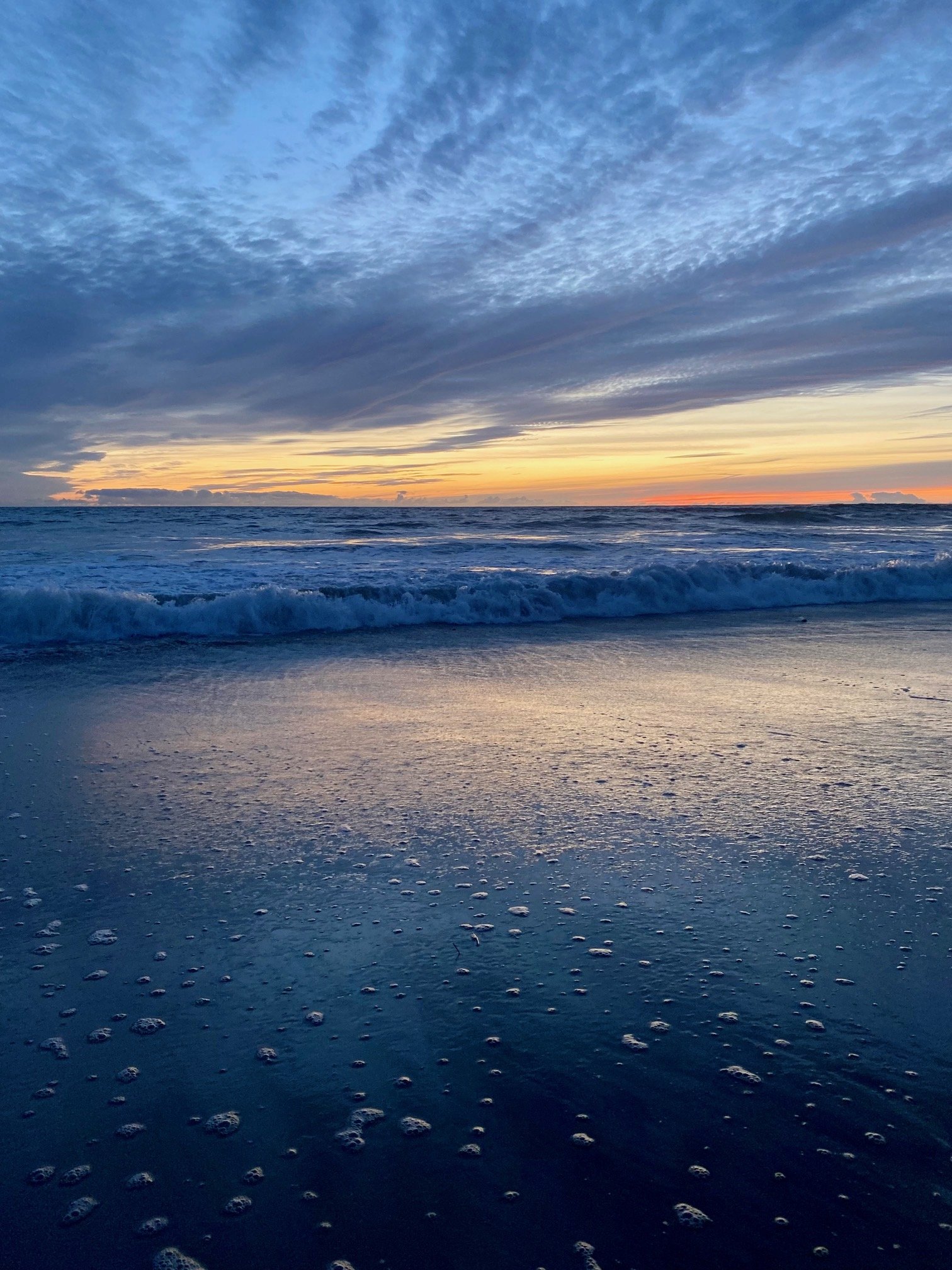 Sunset over the ocean with clouds and waves, wet sandy beach with foam and reflections.