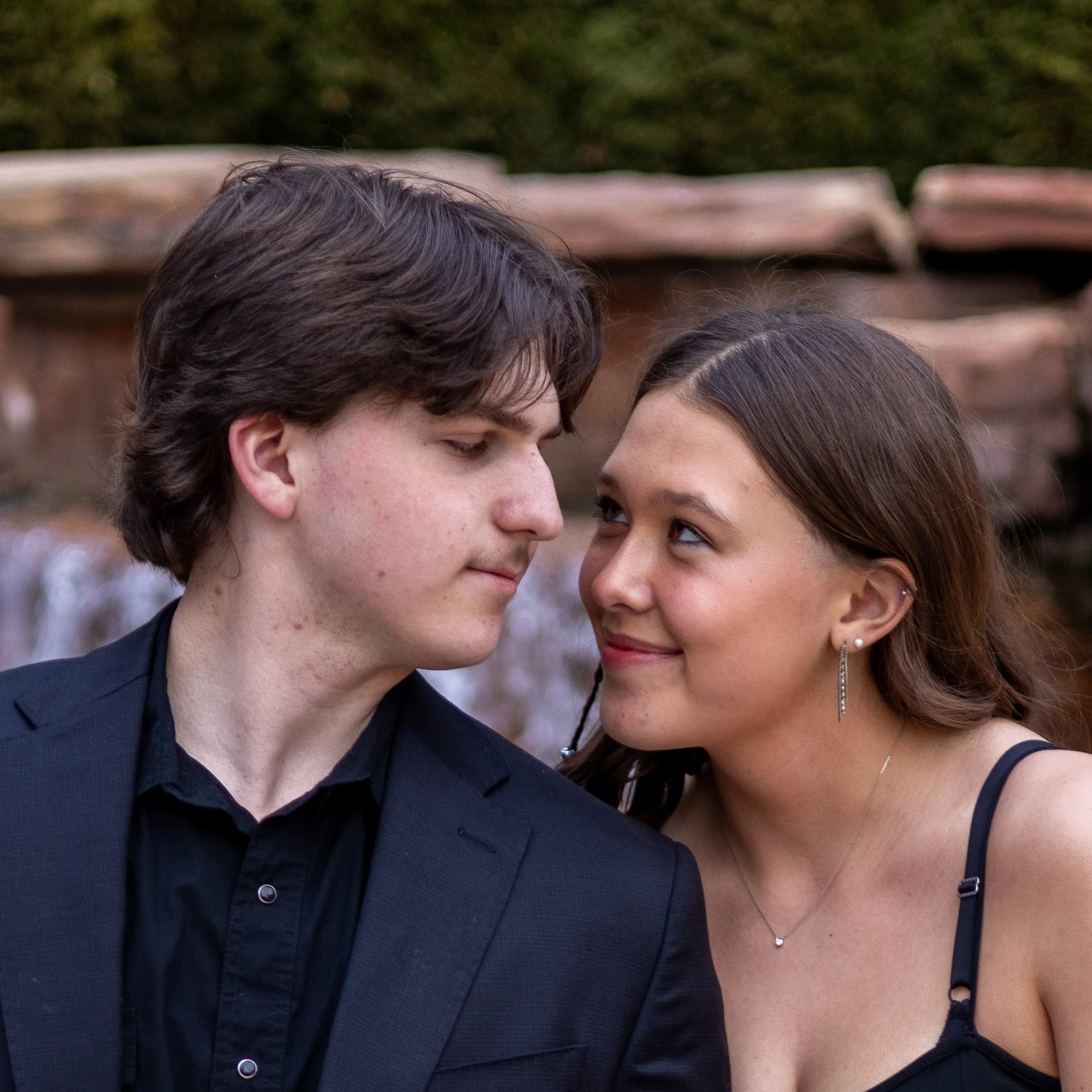 A young man and woman look into each other's eyes during an outdoor event. The man has dark hair and wears a black suit, while the woman has brown hair in loose waves and wears a black dress with thin straps, earrings, and a necklace. They are close 