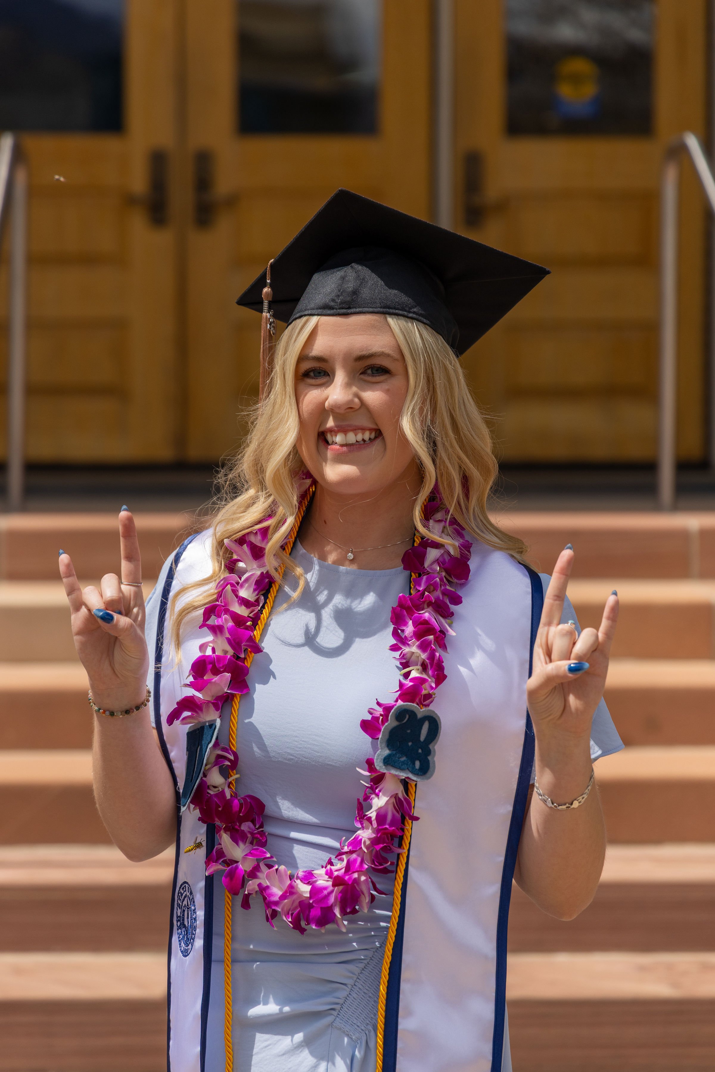 A young woman wearing a graduation cap and gown, adorned with a pink and purple lei, smiling and making peace signs with both hands in front of wooden doors and steps.