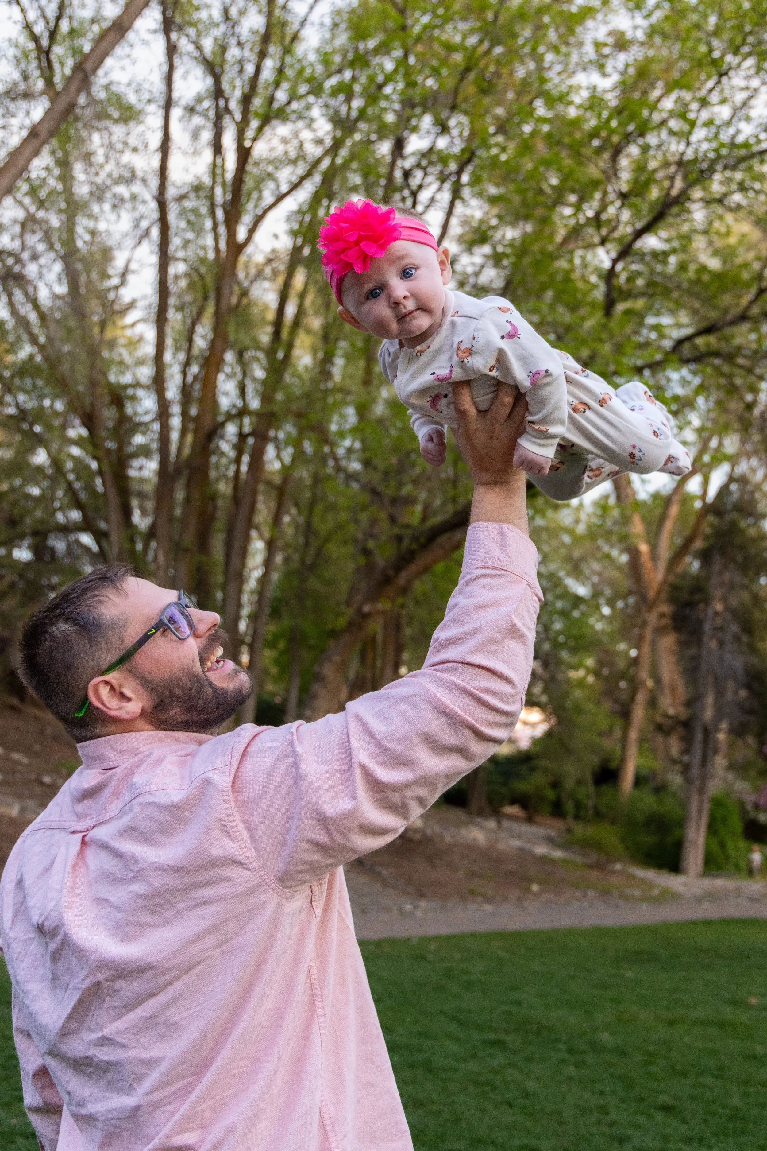 A man with glasses and a beard lifting a baby girl wearing a pink headband with a flower and a pajamas with flamingos, outdoors in a park with trees in the background. Taken by Tucker Davis at the American Fork, Utah, amphitheater.