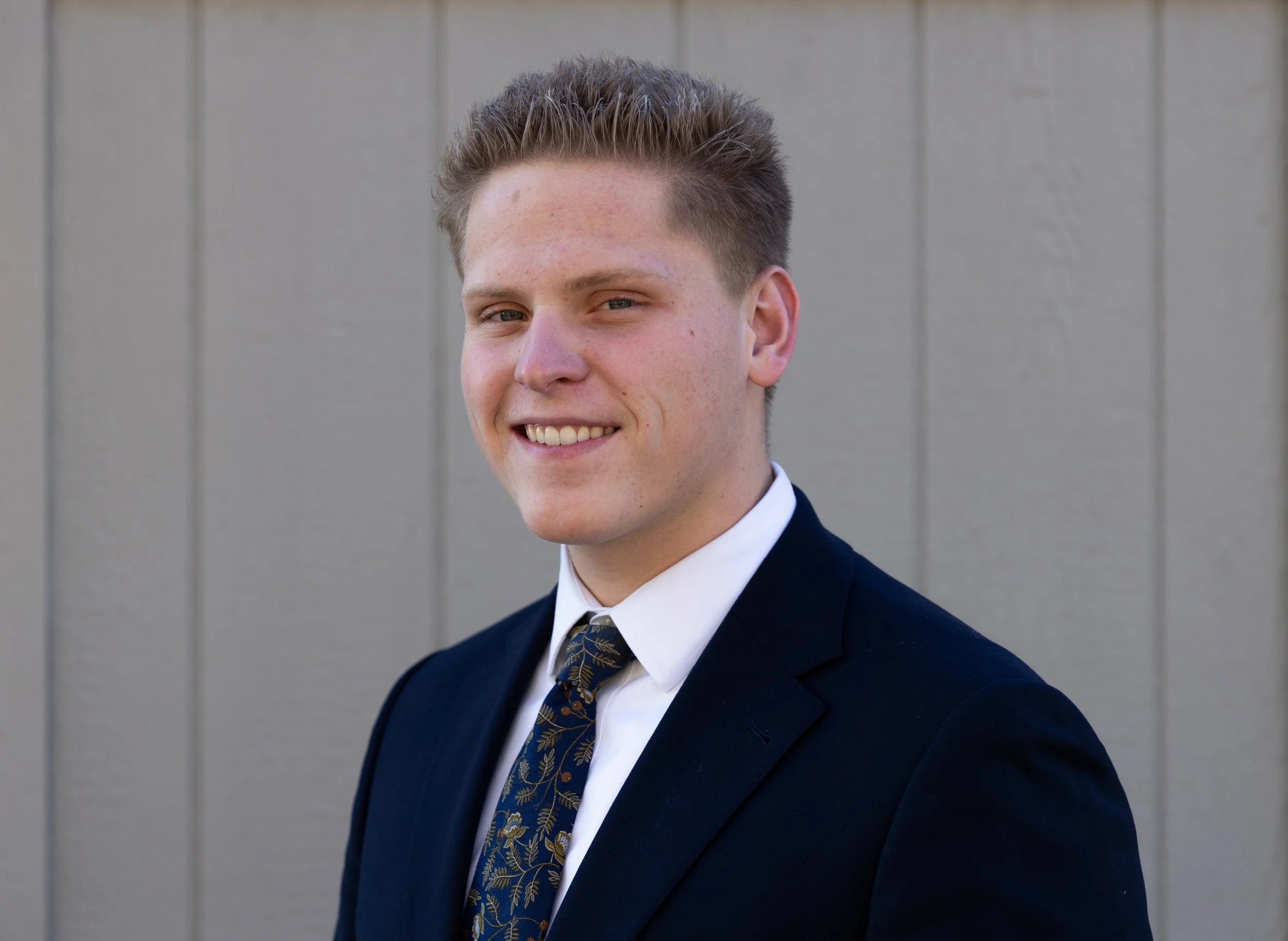 Portrait of a young man with light skin, short light brown hair, wearing a dark suit and a white shirt, standing against a gray wooden background.