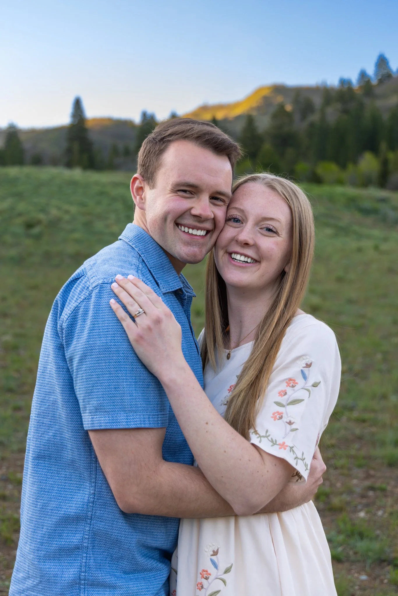 A smiling young man and woman embracing outdoors with green hills and trees in the background.