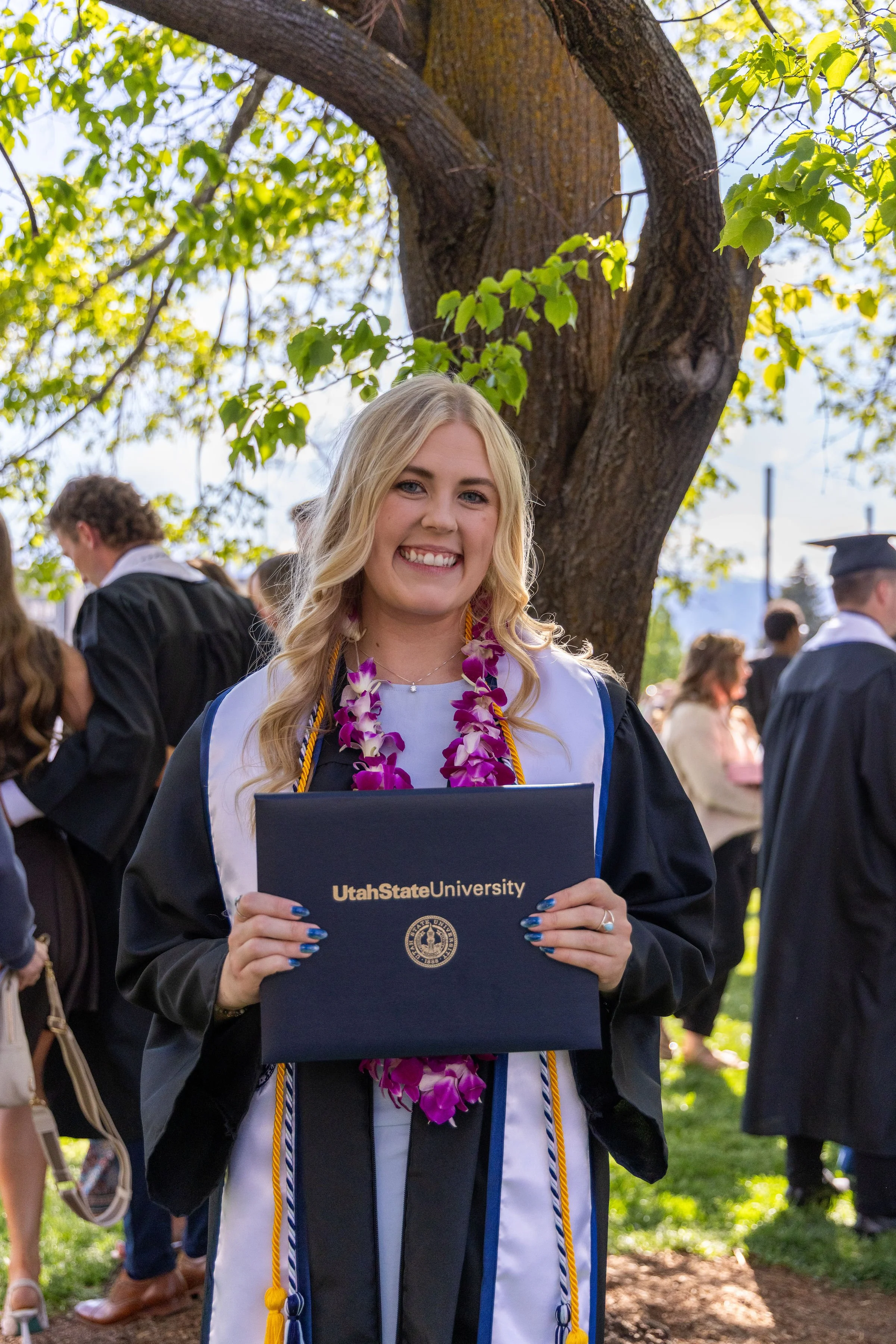 Young woman with blonde hair smiling at graduation ceremony, holding a Utah State University diploma, with people and a large tree in the background.