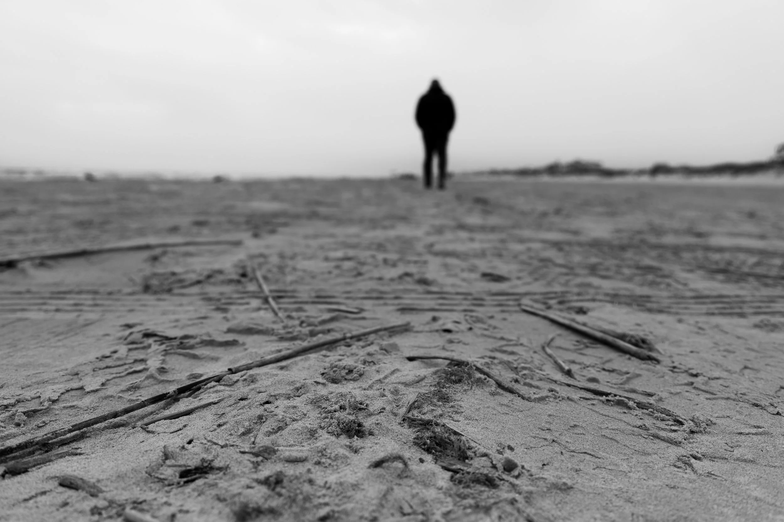 A black and white photo of a person walking away on a beach with sand and scattered sticks, viewed from ground level.