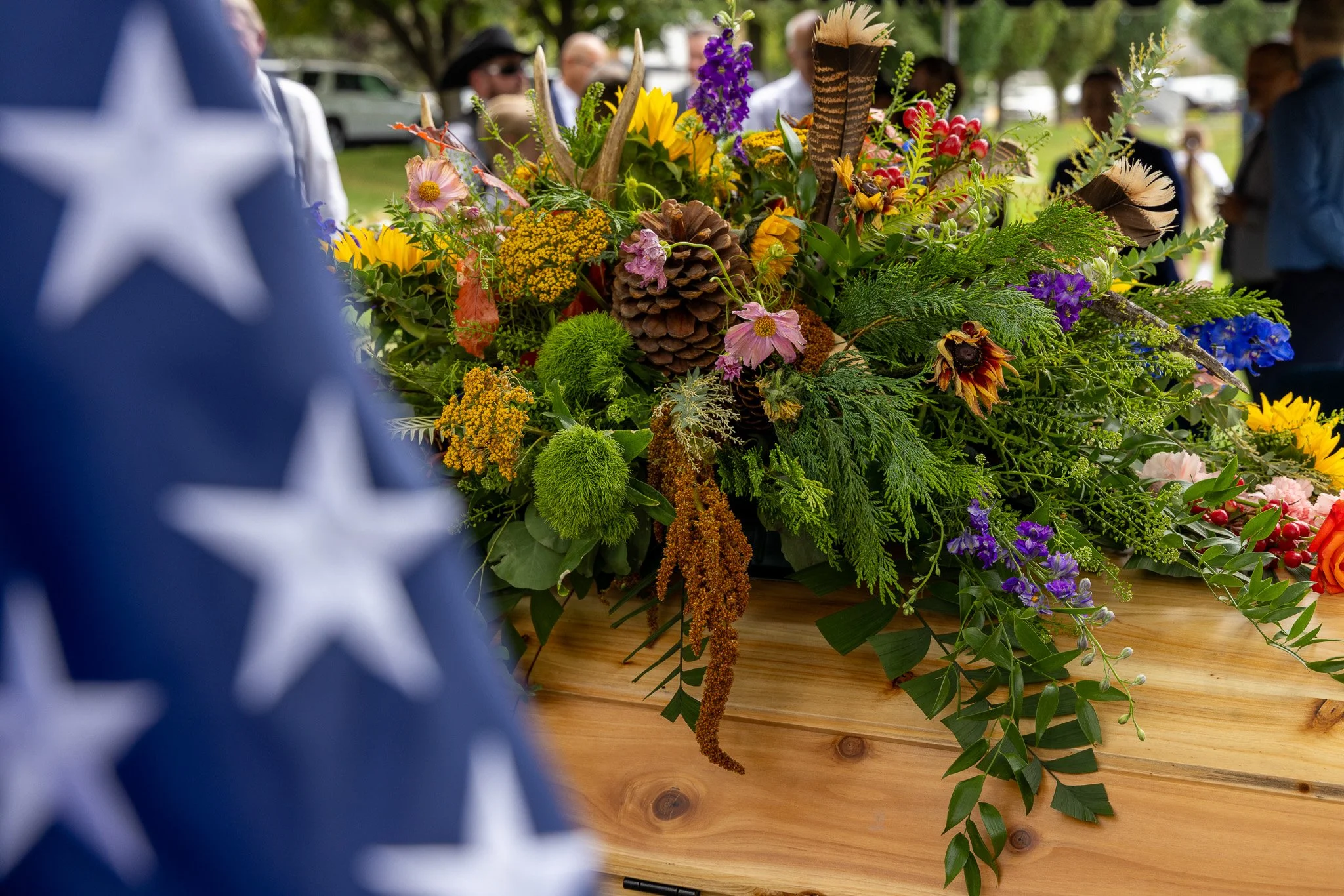 A colorful flower arrangement on a wooden surface, with an American flag in the foreground and people in the background.