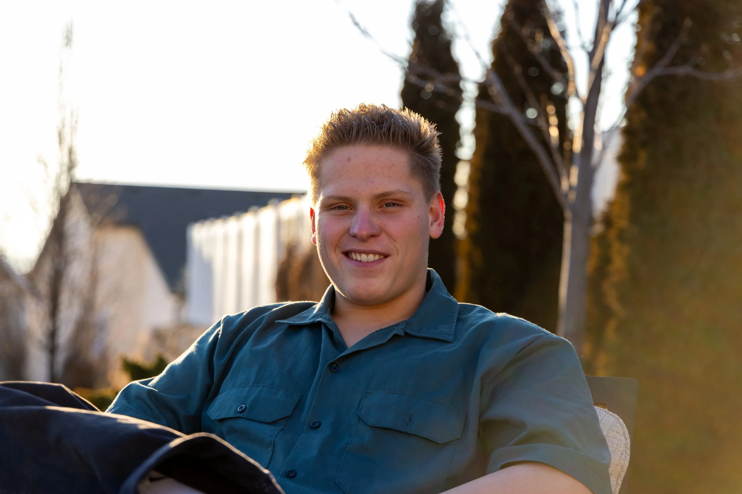 A young man with short, light brown hair smiling at the camera outdoors during sunset, sitting in a chair with trees and a house in the background.