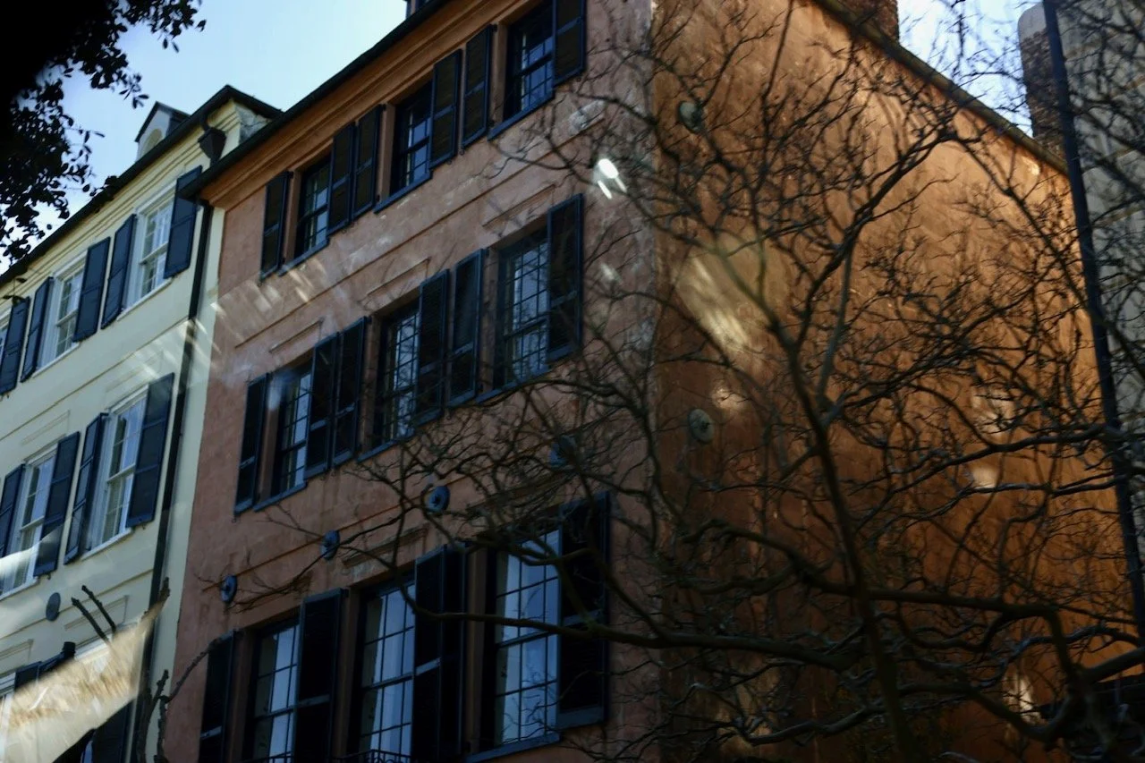 Reflection of a building with orange coloring, black window shutters, and leafless tree in the glass window.