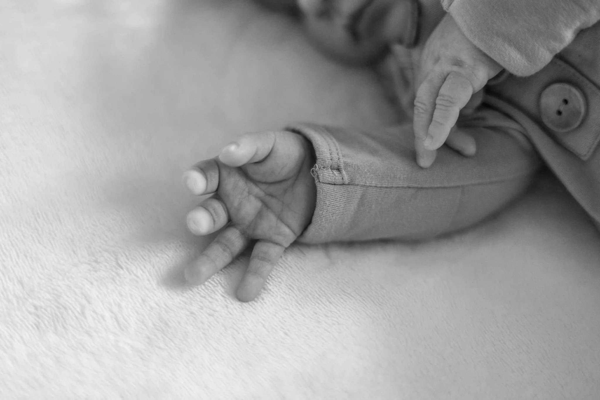 A close-up black and white photo of a child's hand with fingers intertwined with an adult's hand, resting on a textured surface.