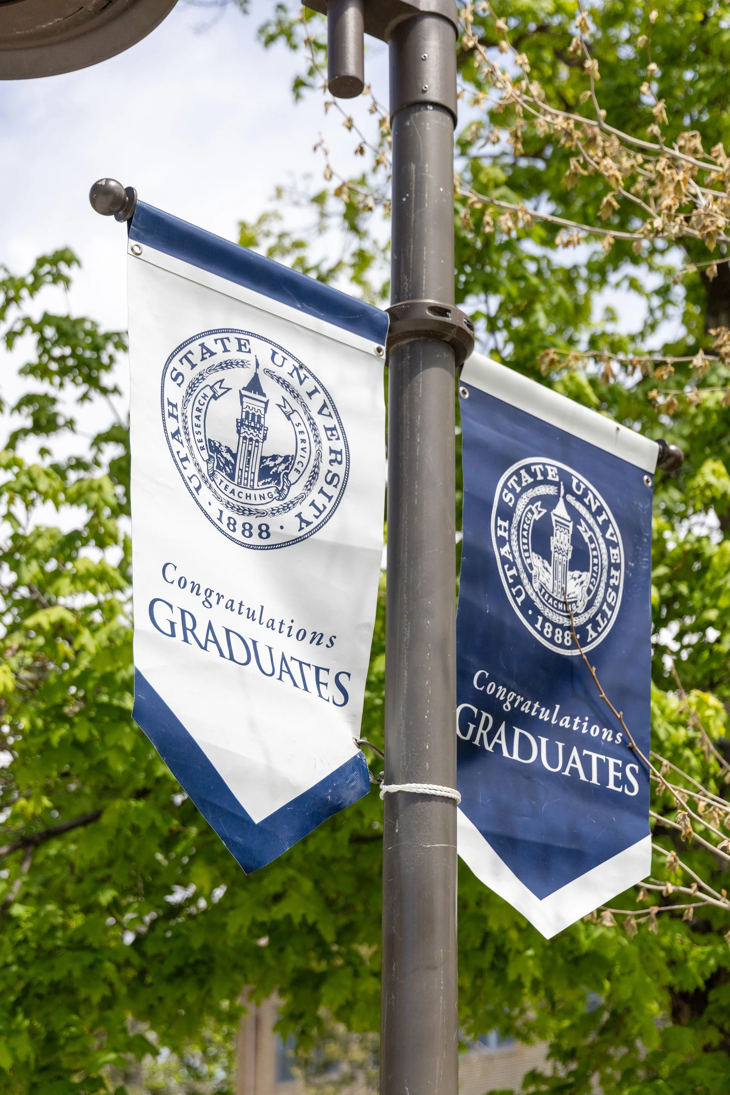 Two banners attached to a gray pole with a tree with green leaves in background. The banners have a circular emblem with a tower and the text 'The State University 1888' and 'Research, Teaching, Service'. The banner in front says 'Congratulations Gra