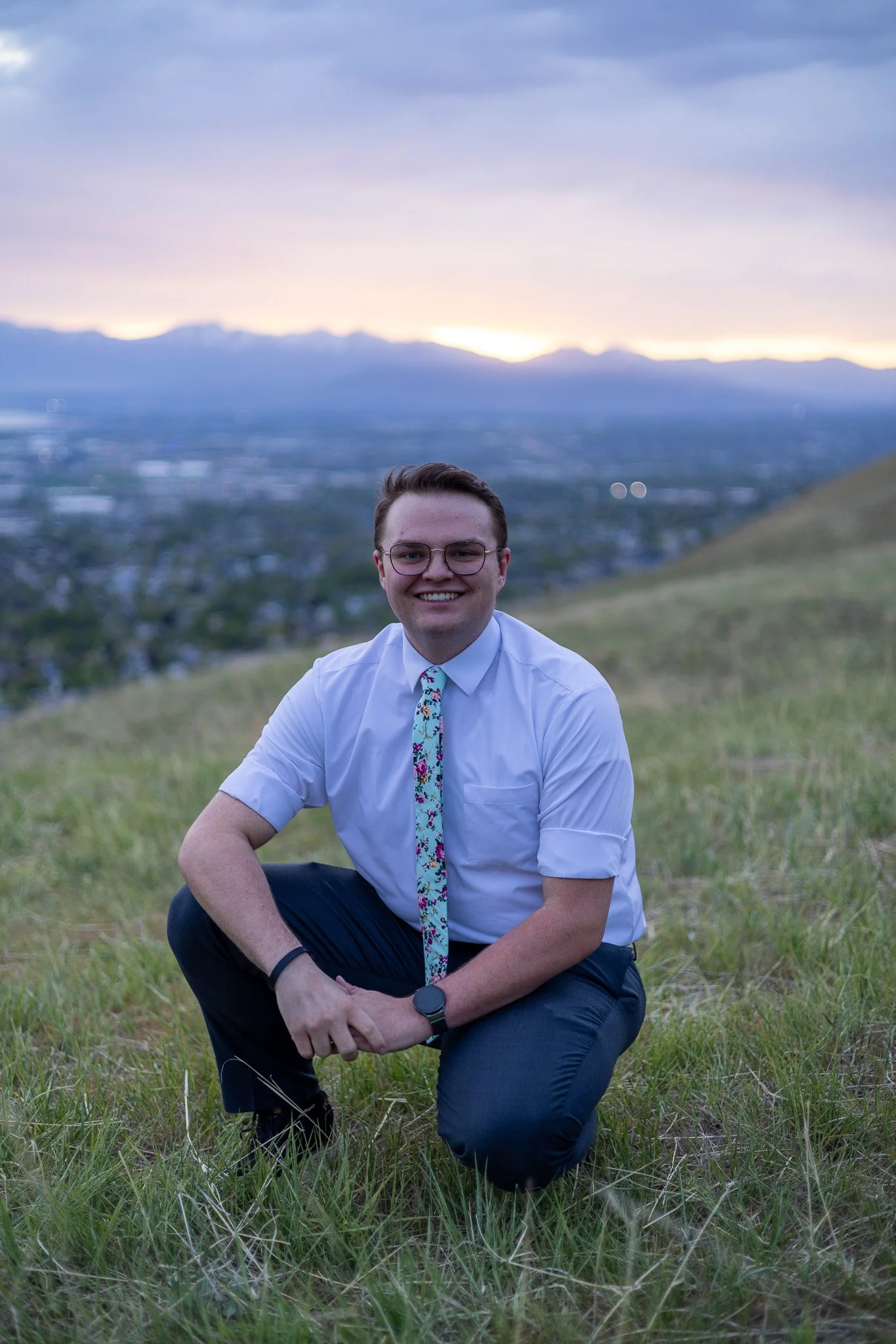 A smiling man in a white shirt, colorful tie, and glasses kneels on grass with a city and mountain range in the background during sunset.