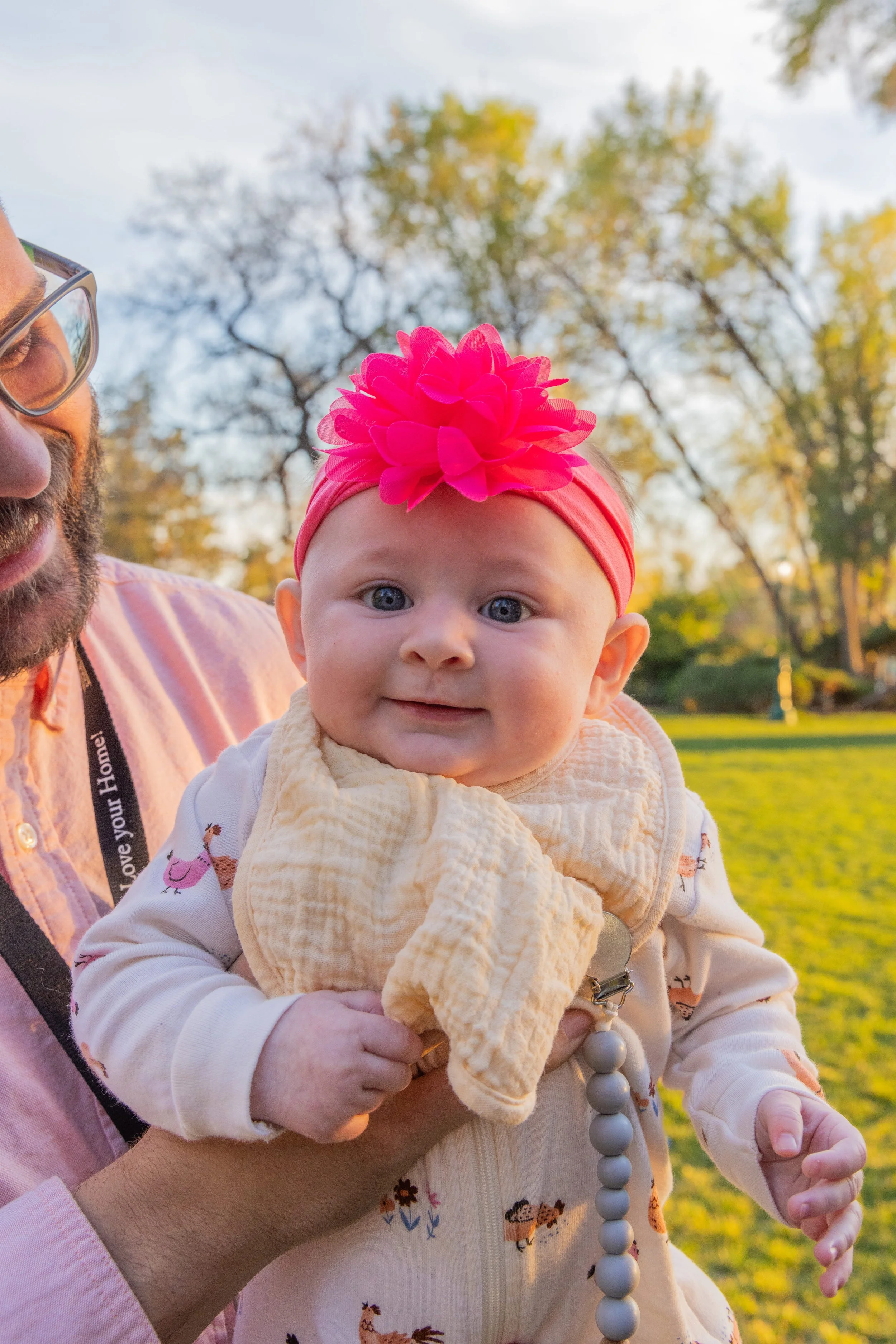 A smiling baby girl with bright blue eyes wearing a pink headband with a large pink flower, held by an adult man in a park with trees and green grass in the background during sunset. Taken by Tucker Davis at the American Fork, Utah, amphitheater.