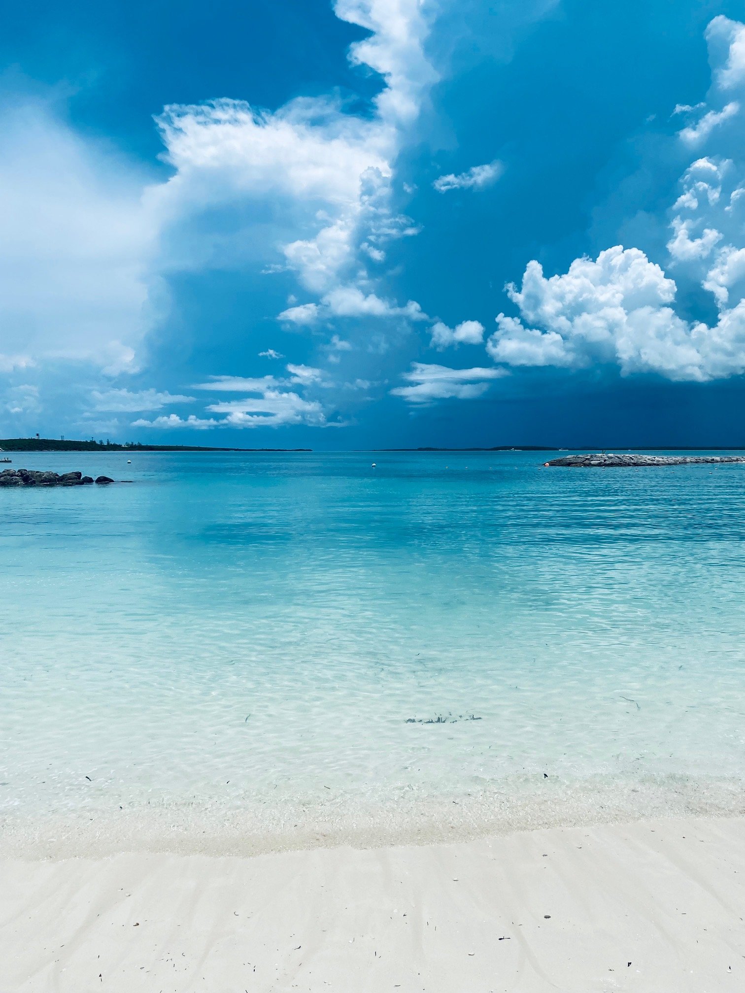 A clear blue sky with white clouds over a calm ocean with light turquoise water and white sandy beach in the foreground.