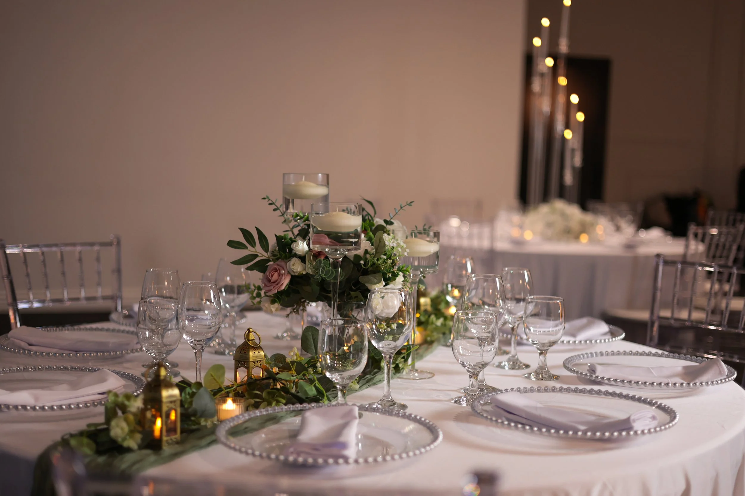 Wedding reception table decorated with floral arrangements, candles in glass holders, and white napkins on clear plates, with empty wine glasses and silver chairs.