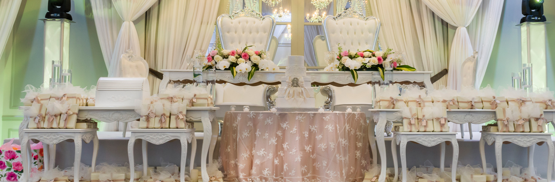 A wedding reception setup with a decorated table and chairs, floral arrangements, a tiered cake, and a backdrop of draped white curtains and chandeliers.