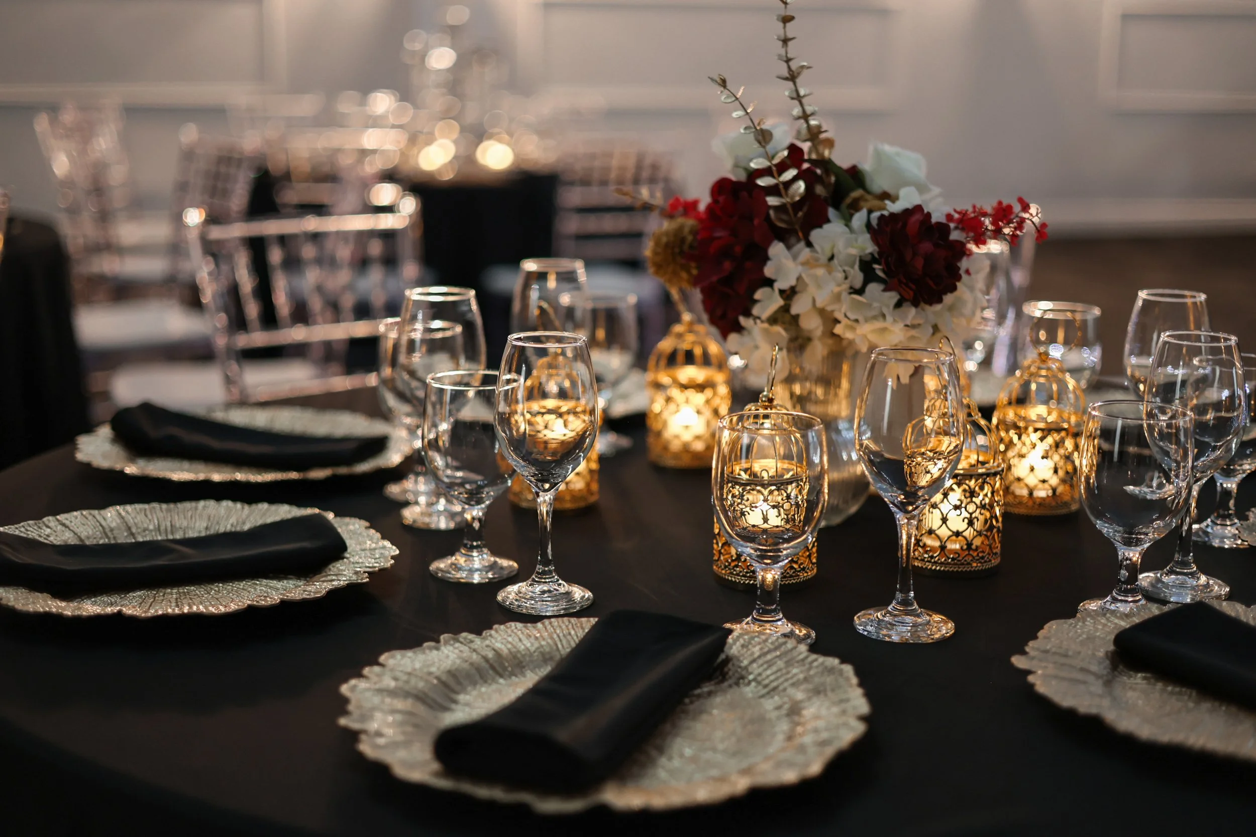 Elegant table setting with glassware, black napkins, decorative plates, and a floral centerpiece amid candles and soft lighting at a formal event or dinner party.
