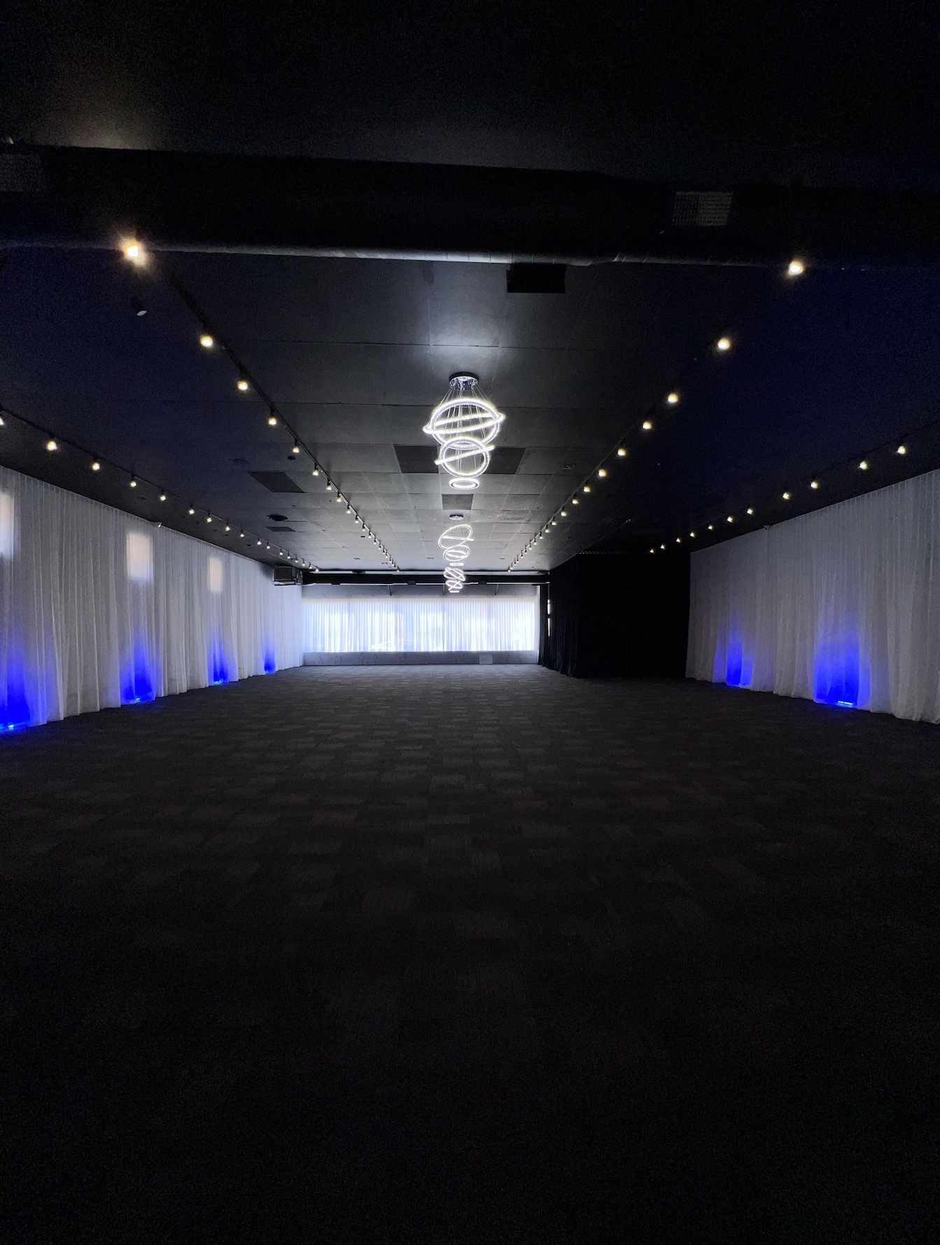 Empty event space or ballroom with black ceiling, white curtains with blue lighting accents, and modern circular chandelier, illuminated by small ceiling lights.