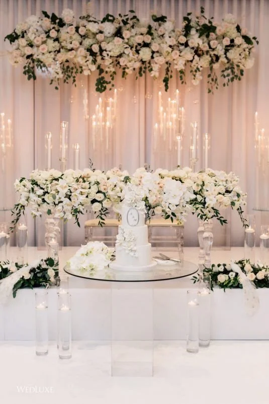 Elegant wedding reception setup featuring a tiered white cake with floral decorations on a glass table, surrounded by white floral arrangements and candles. A floral arch with roses and greenery hangs overhead, with soft lighting and draped curtains 