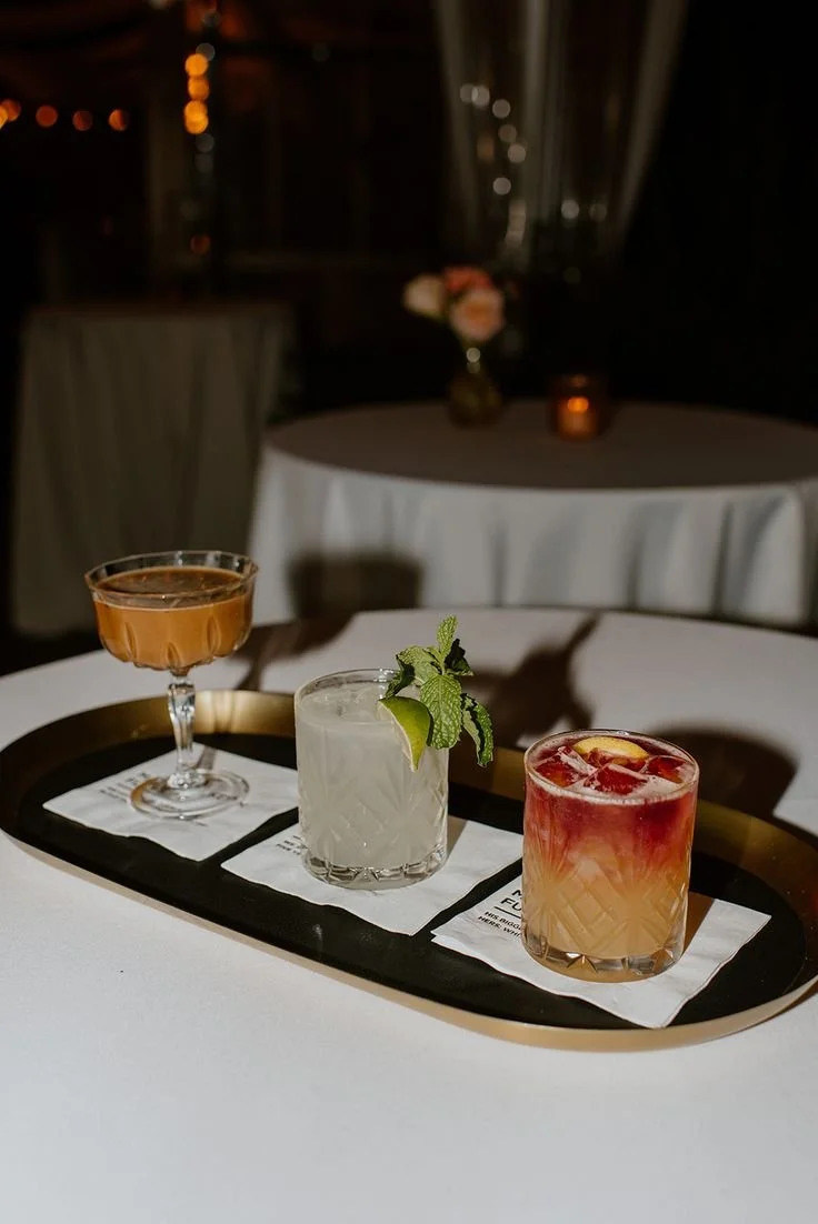 Three cocktails on a black tray, one with a brown liquid, one white with mint, and one red with fruit garnish, on a white table in a dimly lit restaurant.