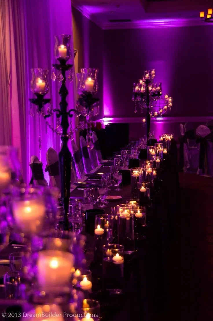 Elegant banquet table decorated with tall candelabras and smaller candles in glass holders, all illuminated with purple lighting.