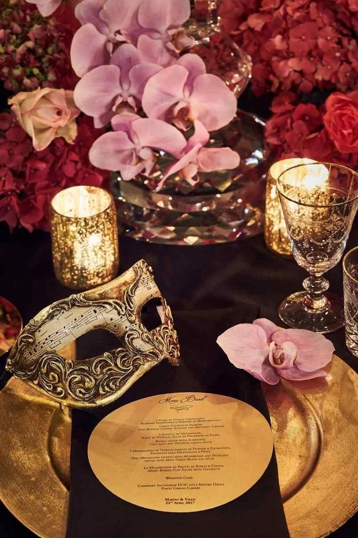 A decorative formal table setting with pink orchids, red and pink flowers, gold candle holders, and a gold Venetian mask with musical notes. There is also a gold charger plate, a printed menu, and glassware.