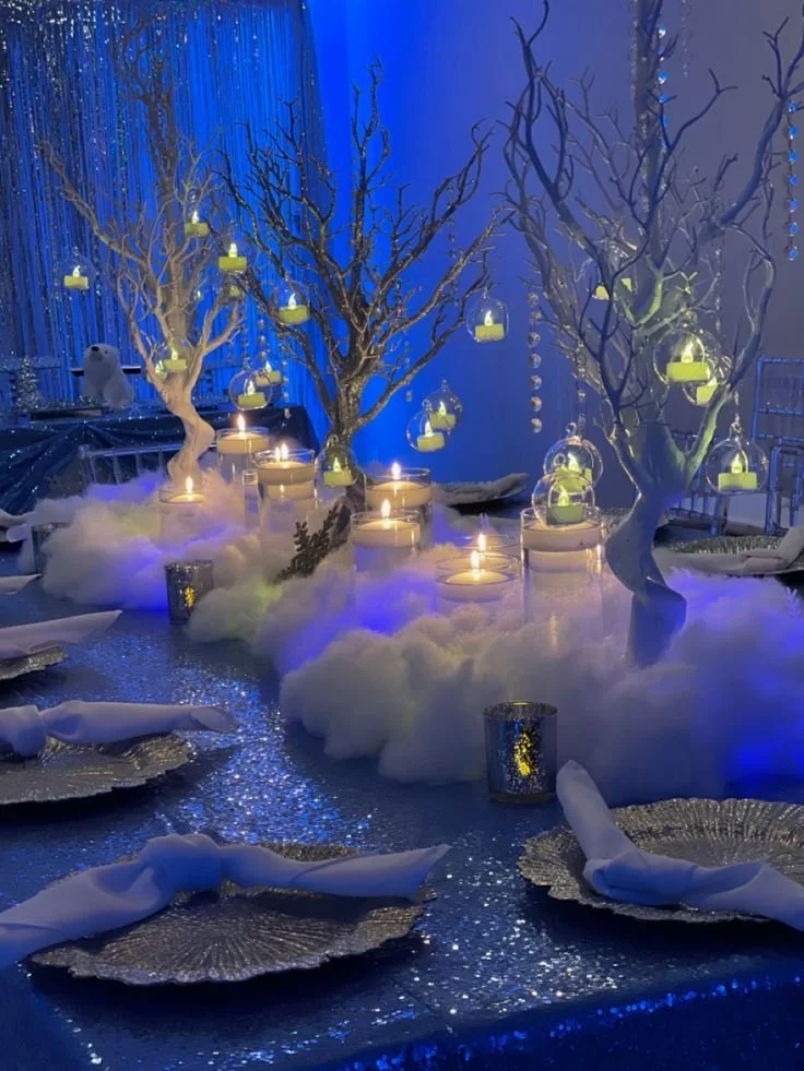 Elegant winter-themed table setting with white cotton resembling snow, decorative white trees with hanging lanterns, candles, and silver accents, illuminated in blue lighting.