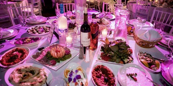 A festive dining table with various dishes, bottles of wine, and candles, illuminated by purple lighting.