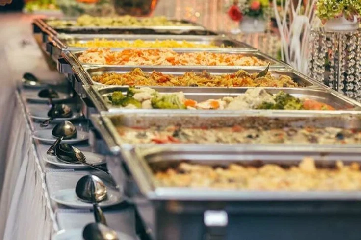 Buffet table with several chafing dishes containing various hot dishes and salads, with serving spoons placed on plates beside each dish, set in a decorated area with flowers.