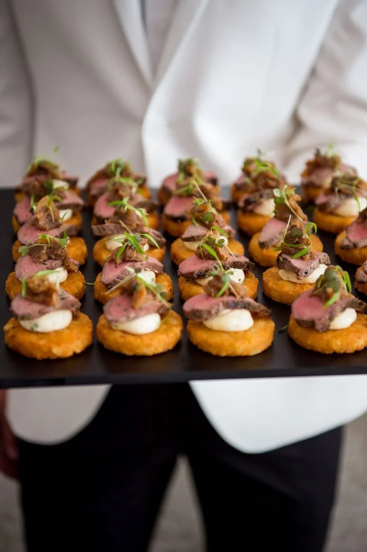 Tray of bite-sized appetizers with slices of beef, dollops of white sauce, and green microgreens on small crispy rounds.