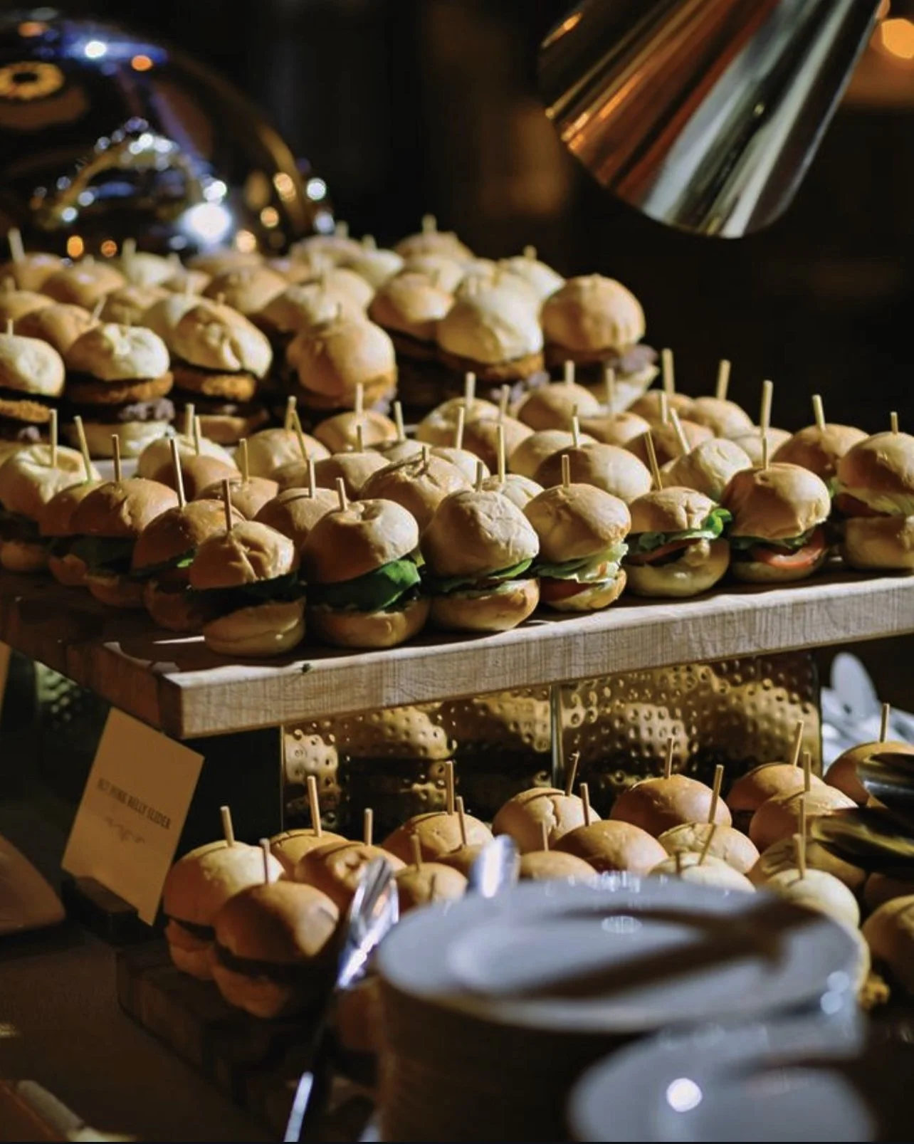 Close-up of a buffet tray with small sliders topped with greens and toothpicks, with a stack of white plates in the foreground.