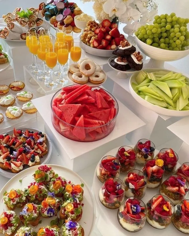 Assorted fruits, desserts, and beverages on a table, including watermelon slices, strawberries, grapes, cookies, mini tarts, juice glasses, and various sweets topped with berries and edible flowers.