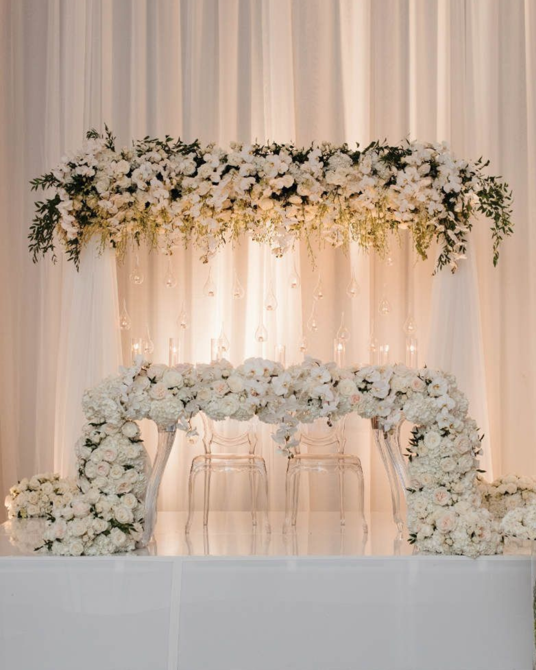 Elegant wedding altar decorated with white and cream flowers, transparent chairs, surrounded by soft white drapery and hanging candles or lanterns.