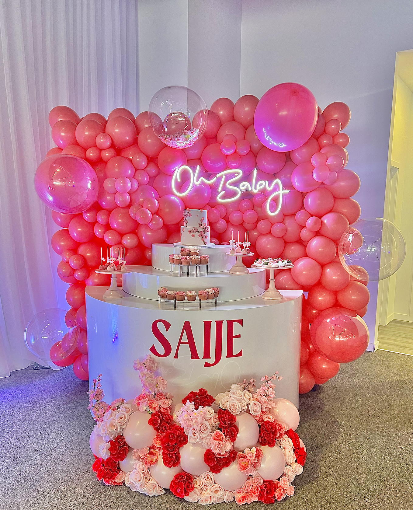 Decorative birthday display with pink balloons, a neon sign reading 'Oh Boy', a tiered cake, cupcakes, and floral arrangements.
